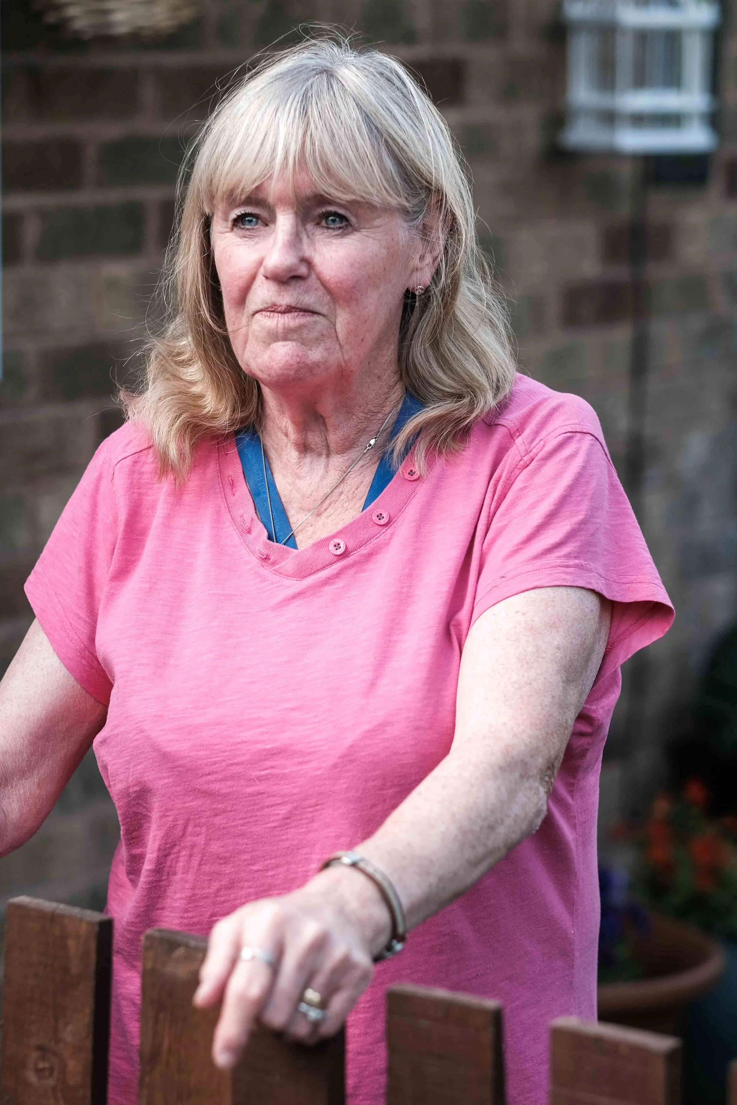 A mature woman with blonde hair wearing a pink T-shirt standing outdoors.