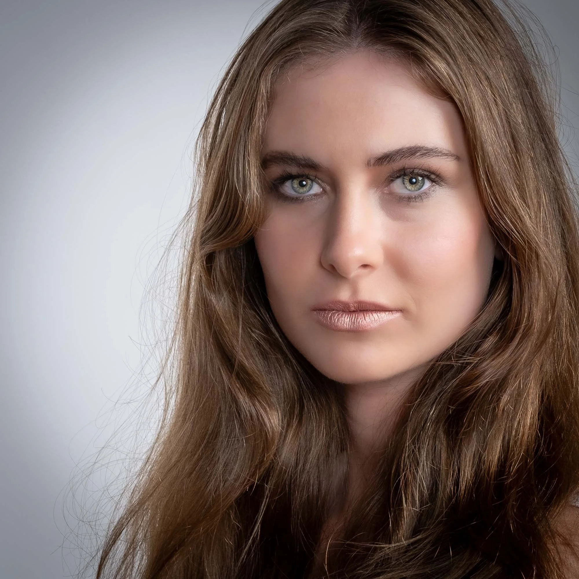 Close-up of a young woman with long, wavy brown hair and blue eyes, facing the camera with a neutral expression.