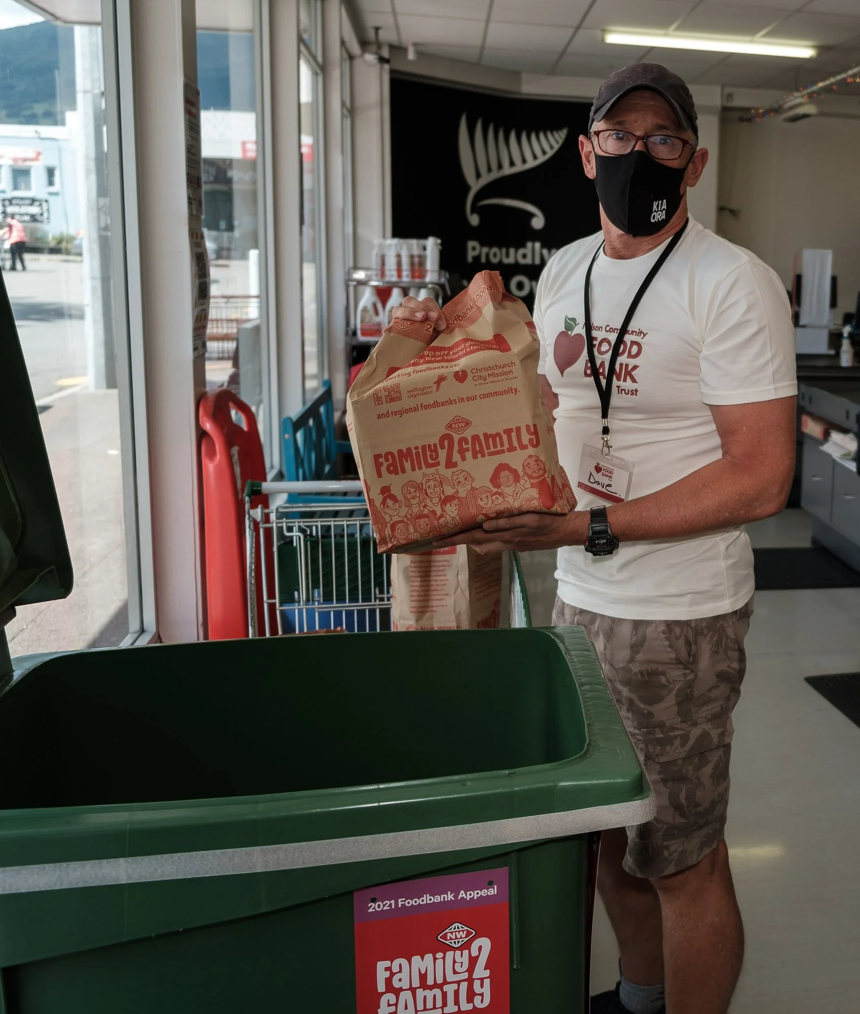 A man wearing glasses, a black face mask, a white T-shirt, and camouflage shorts is dropping a paper bag into a green trash bin inside a store. The paper bag has red and white text and illustrations. The store has large windows, a shopping cart, and 