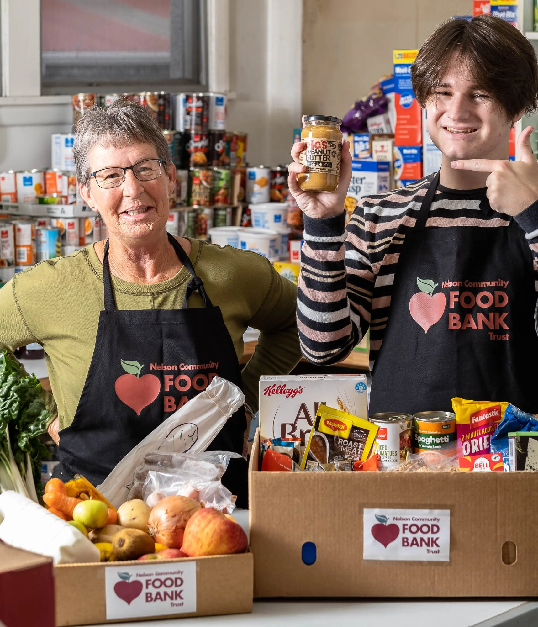 A smiling woman and a young man wearing Nelson Community Food Bank aprons standing behind a table with donated food items including fruits, canned goods, and boxes, at a food bank.