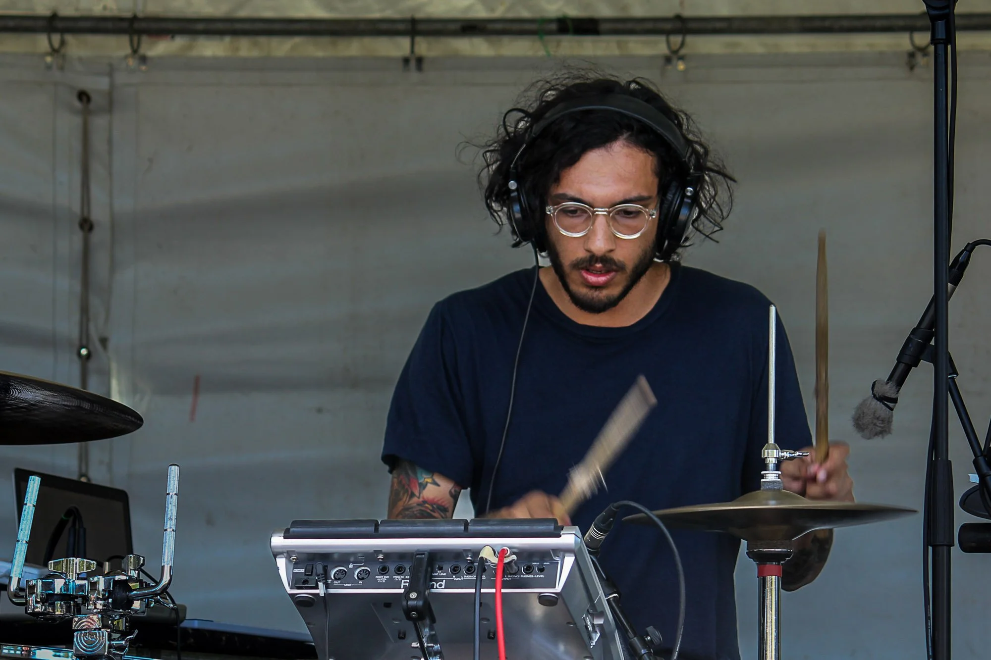 A man with curly hair and glasses playing electronic drums with drumsticks, wearing headphones, surrounded by electronic music equipment.