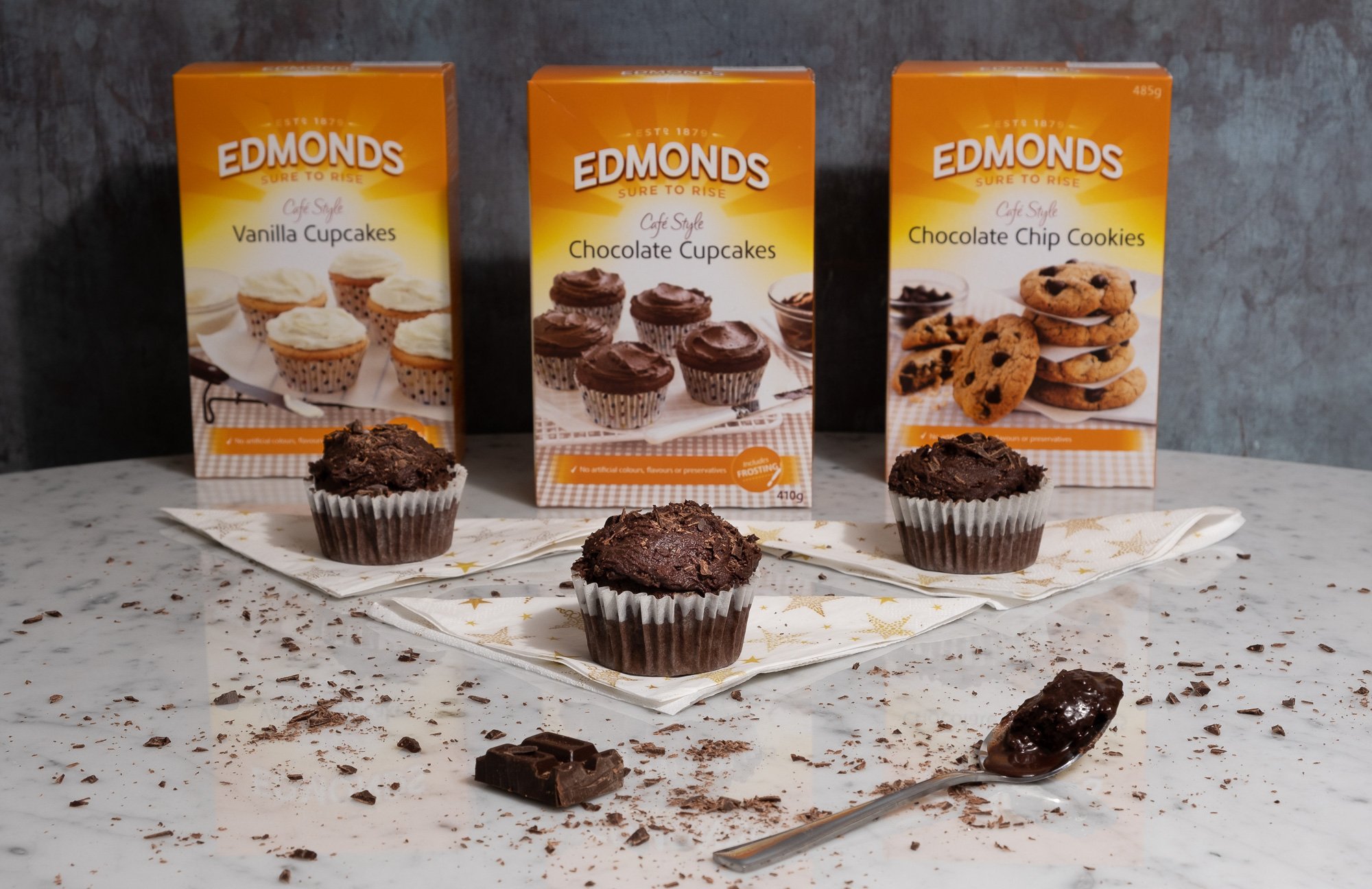 Three chocolate cupcakes on napkins, with boxes of vanilla cupcakes, chocolate cupcakes, and chocolate chip cookies in the background on a marble surface.