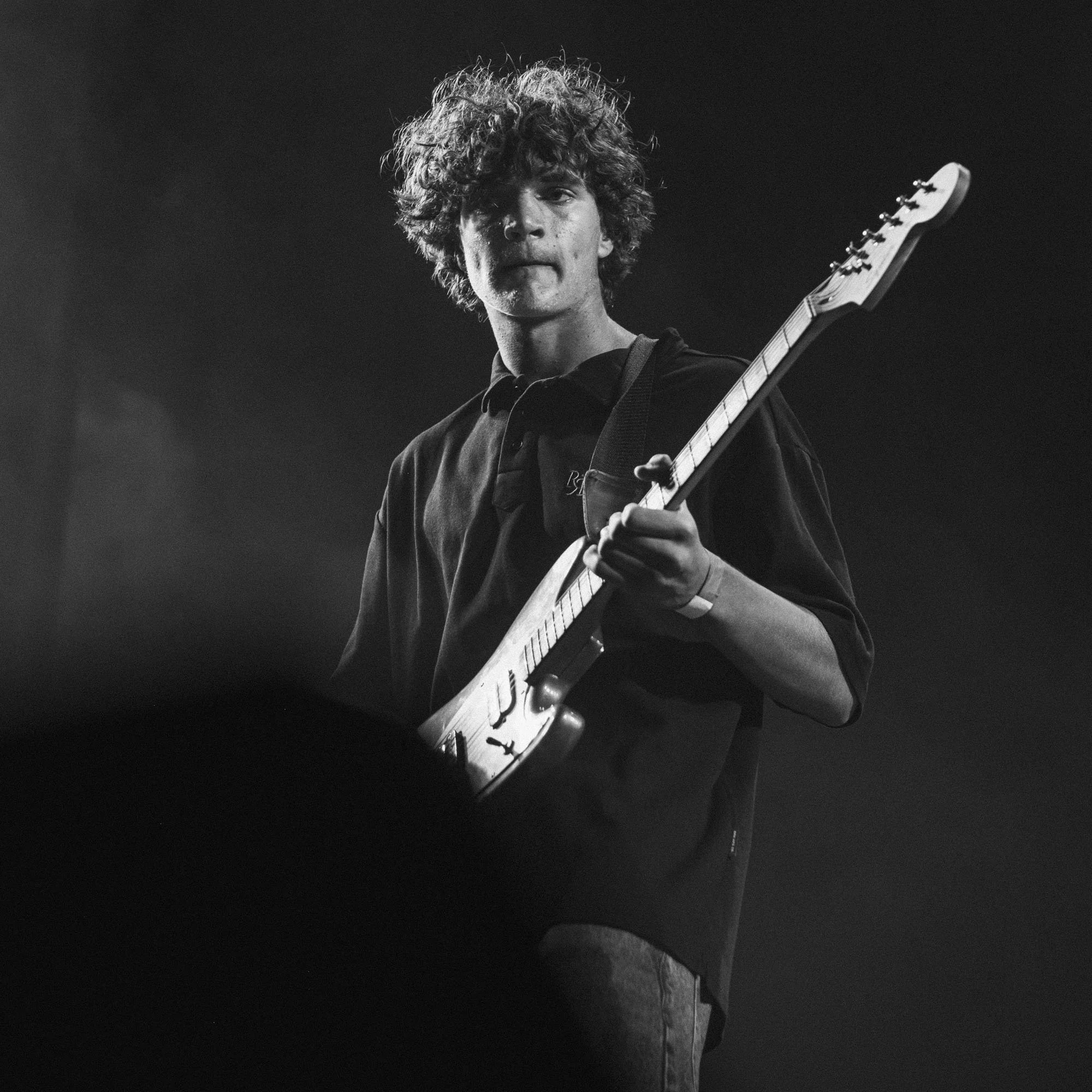 Black and white photo of a young man playing an electric guitar, with curly hair and focused expression.