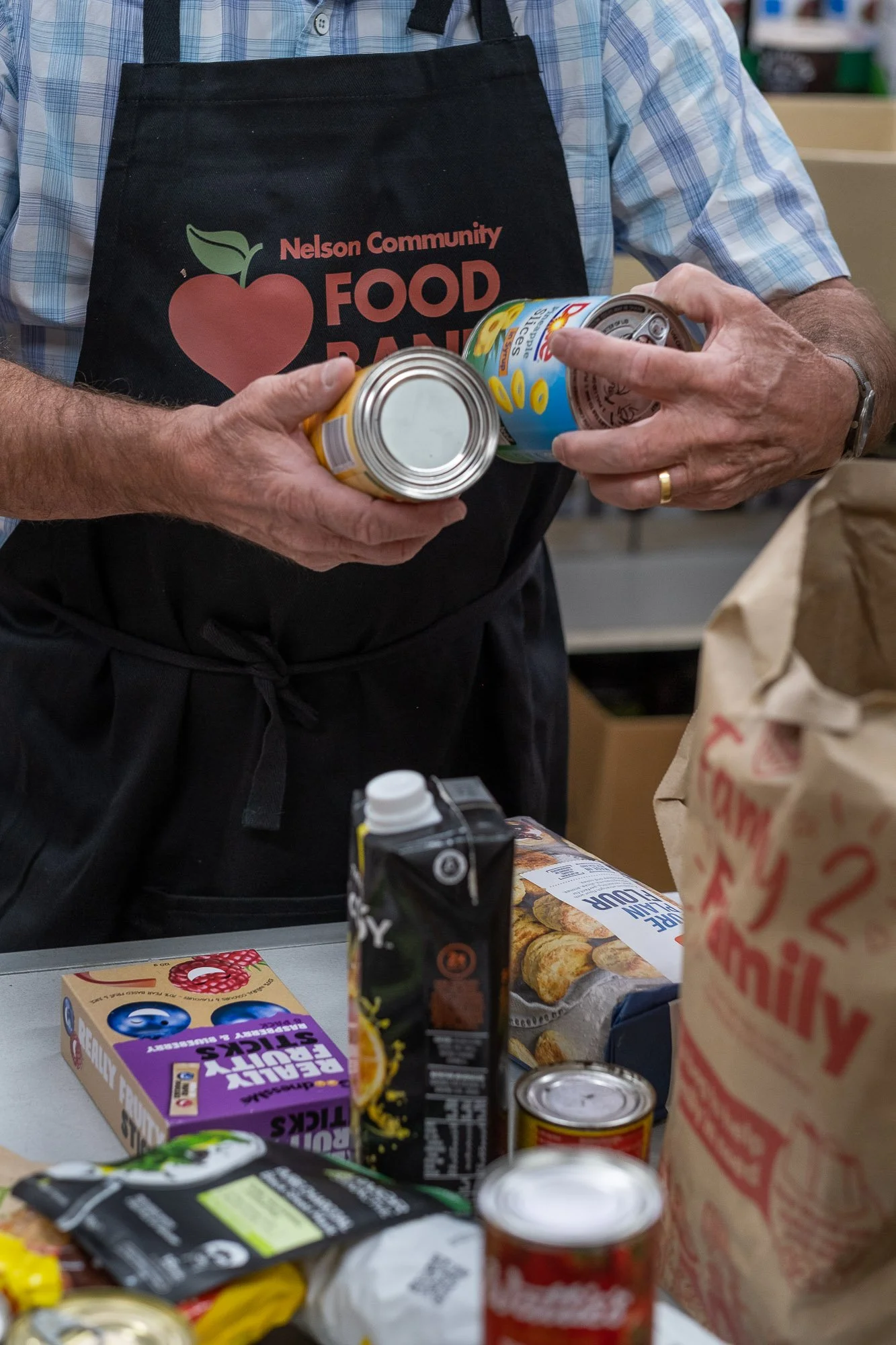 Person wearing a black apron with 'Nelson Community Food Bank' logo, holding canned goods at a food bank.