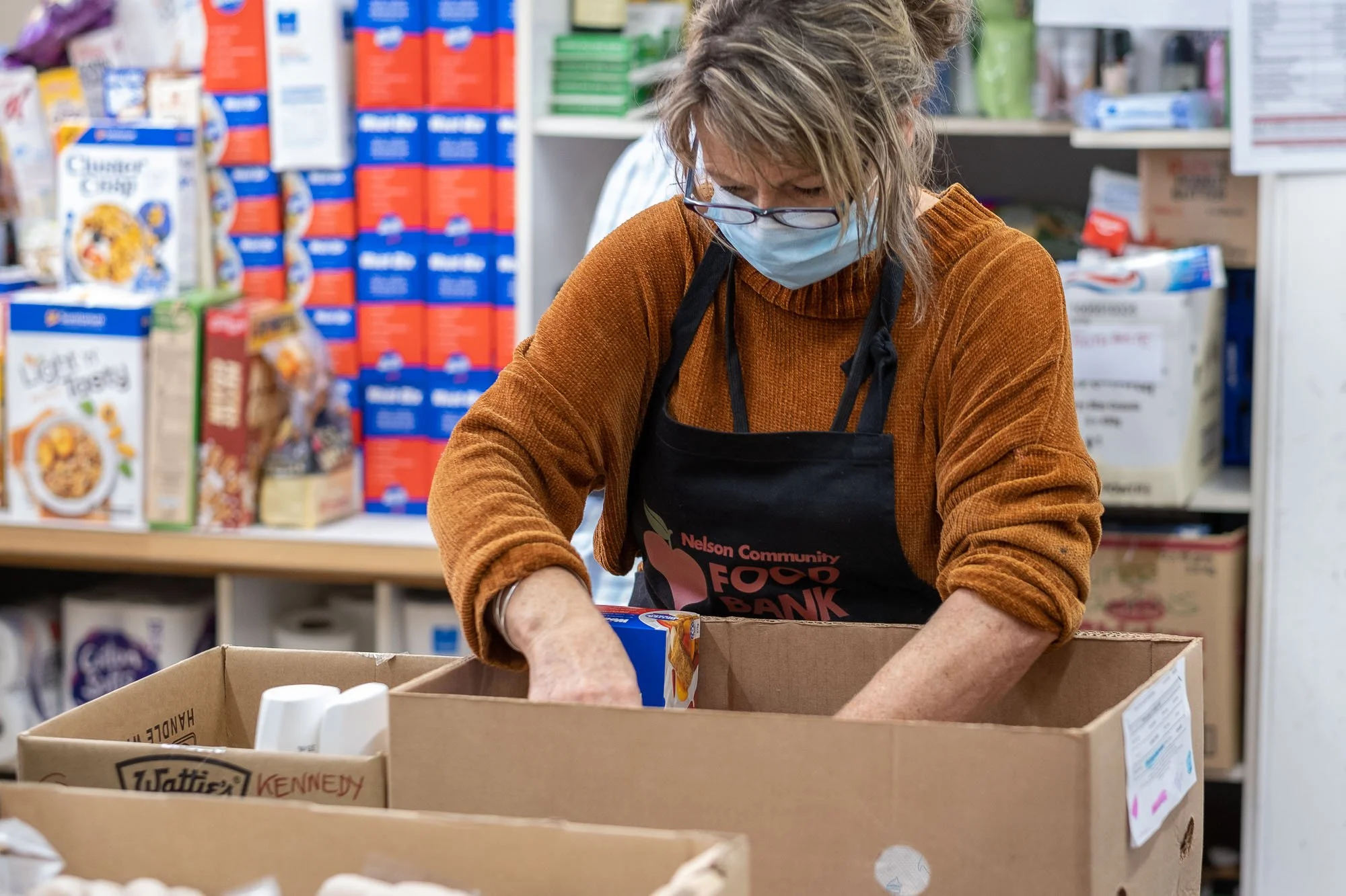 A woman wearing a face mask, glasses, and a brown sweater sorts food in a grocery store or food bank with shelves filled with cereal boxes in the background.