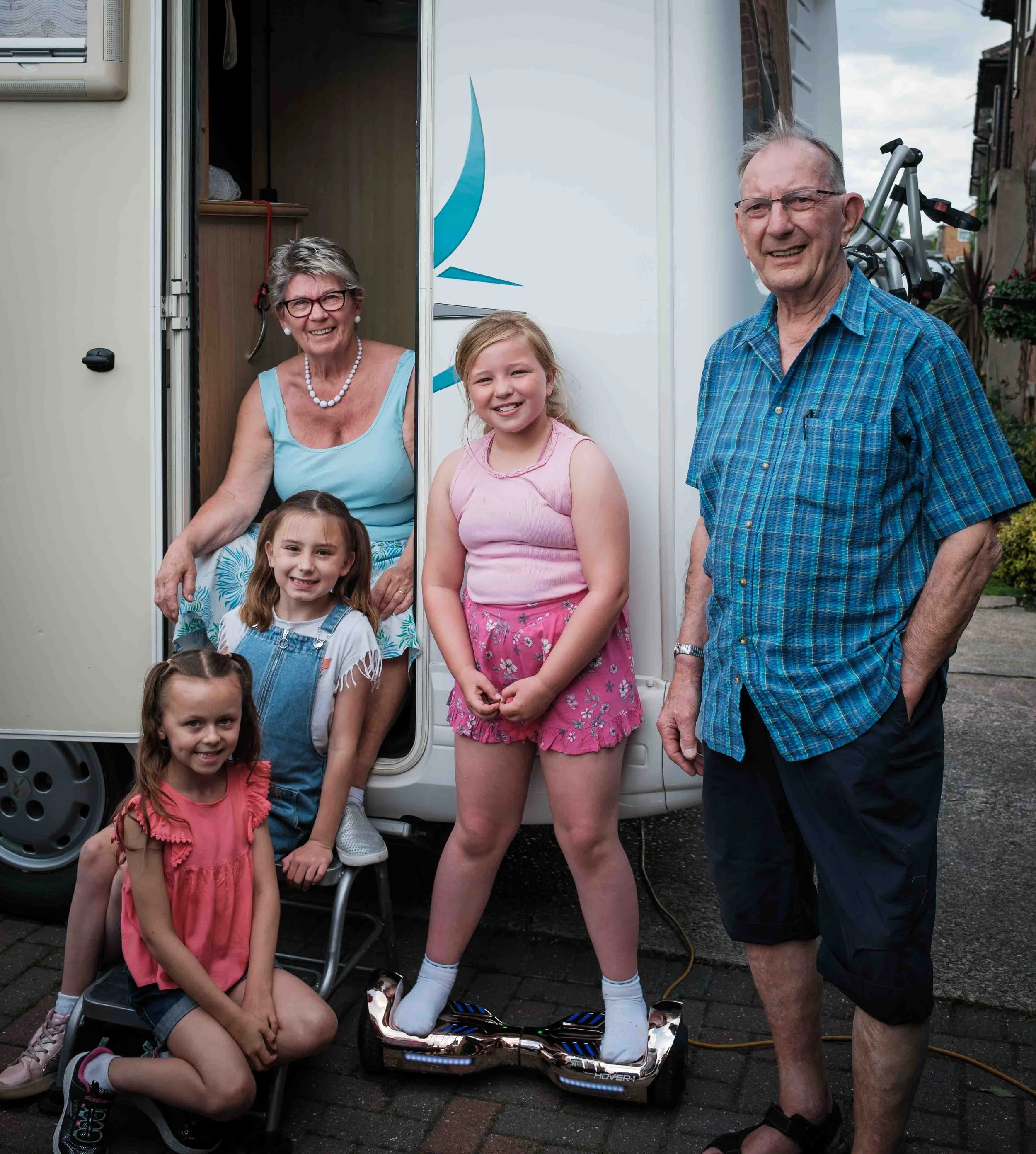 Family outside their campervan, including elderly man and woman with four young girls smiling, one girl standing on a hoverboard.