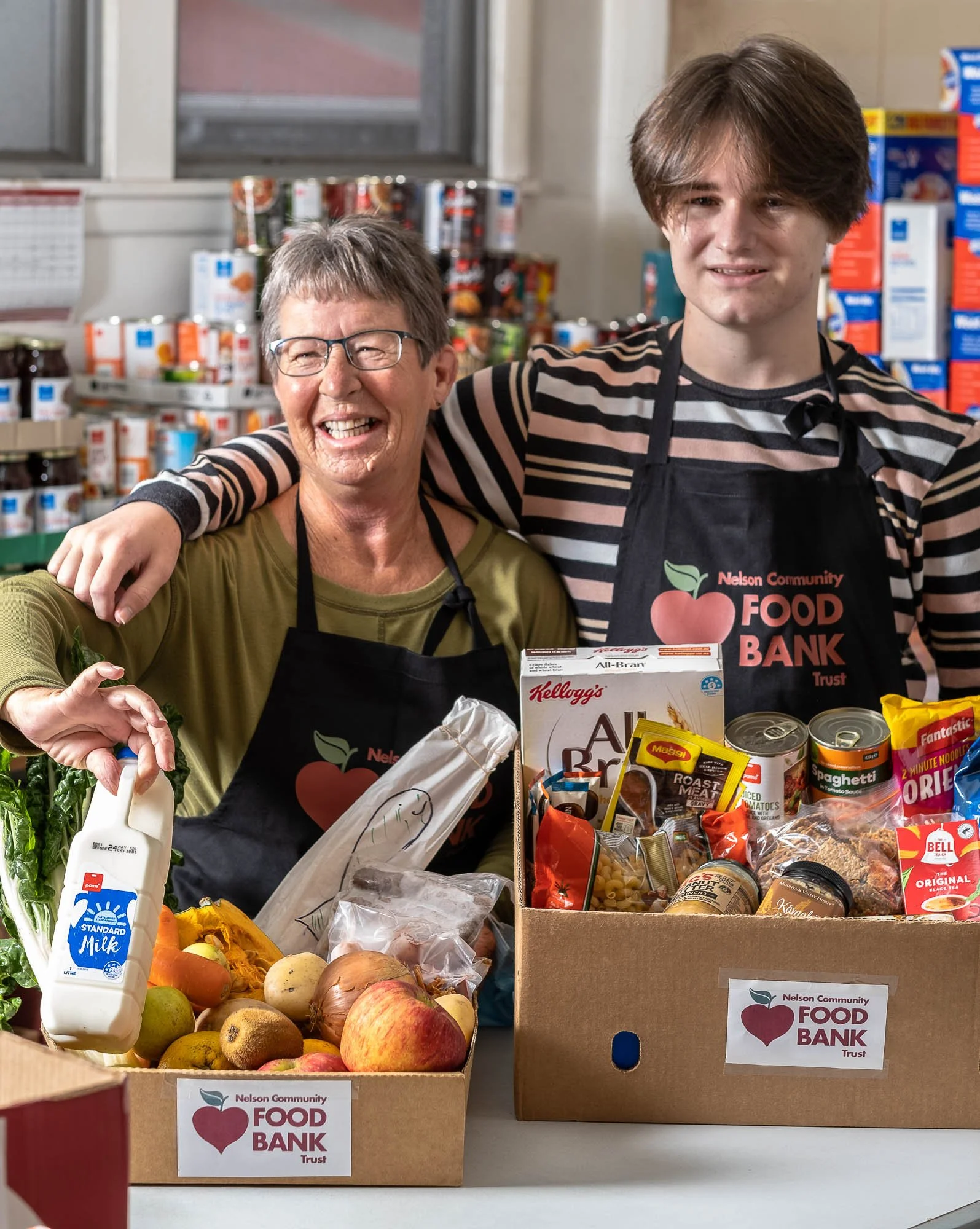 A smiling elderly woman and teenage boy in an aisle near a food bank with boxes of donated groceries, including fruits, vegetables, canned goods, and packaged food.