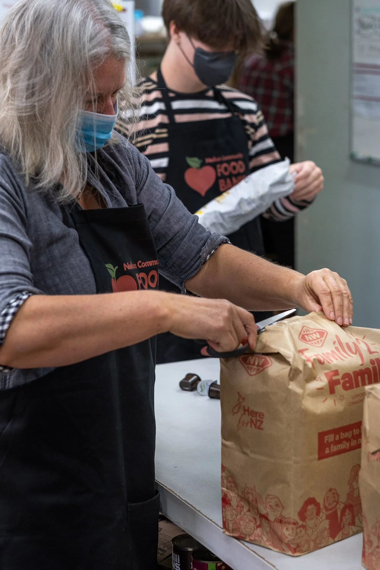 Two people wearing face masks and aprons are packing food into brown paper bags in a room, with canned goods on the table.