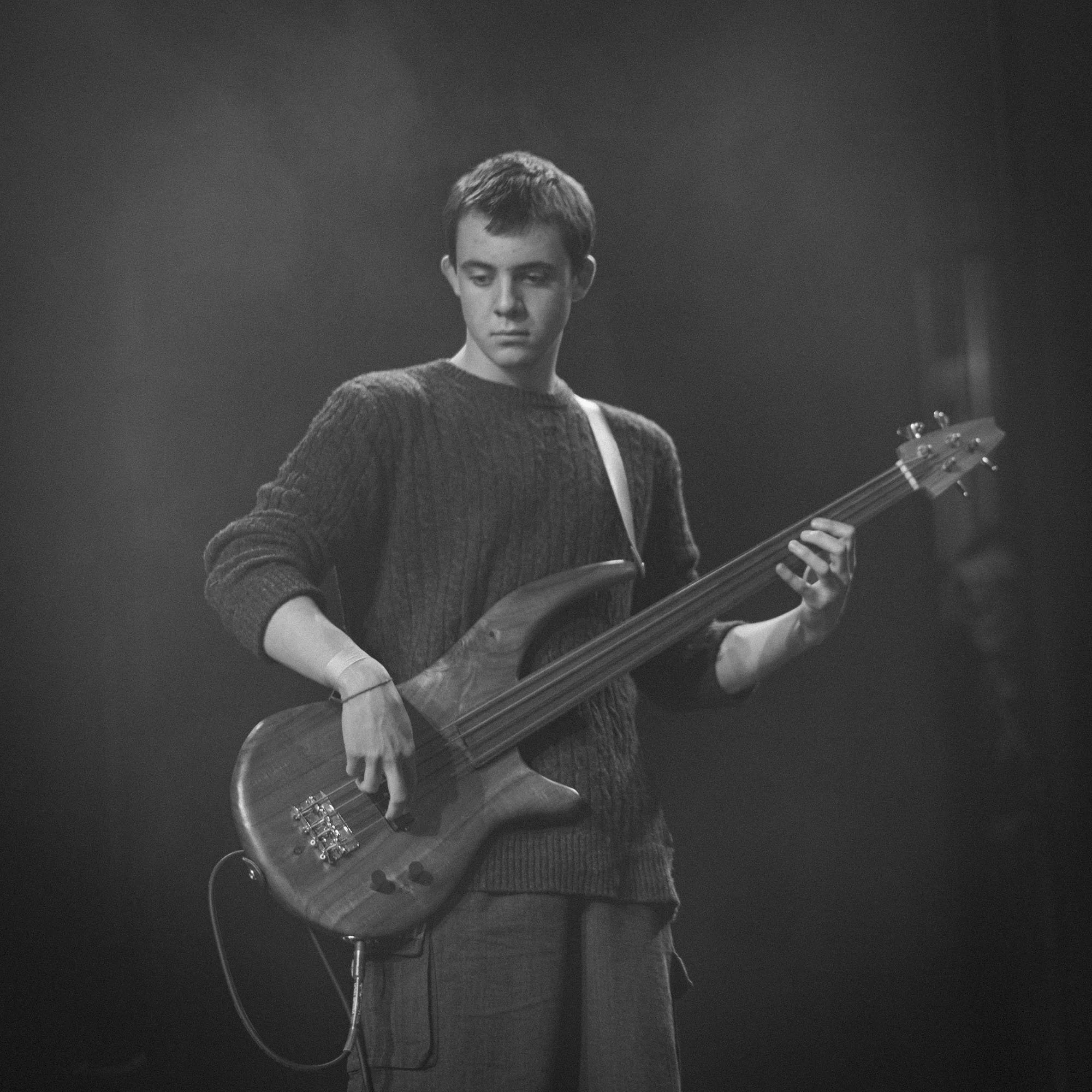 A young man playing an electric bass guitar on stage, wearing a sweater, in black and white, with a focused expression.