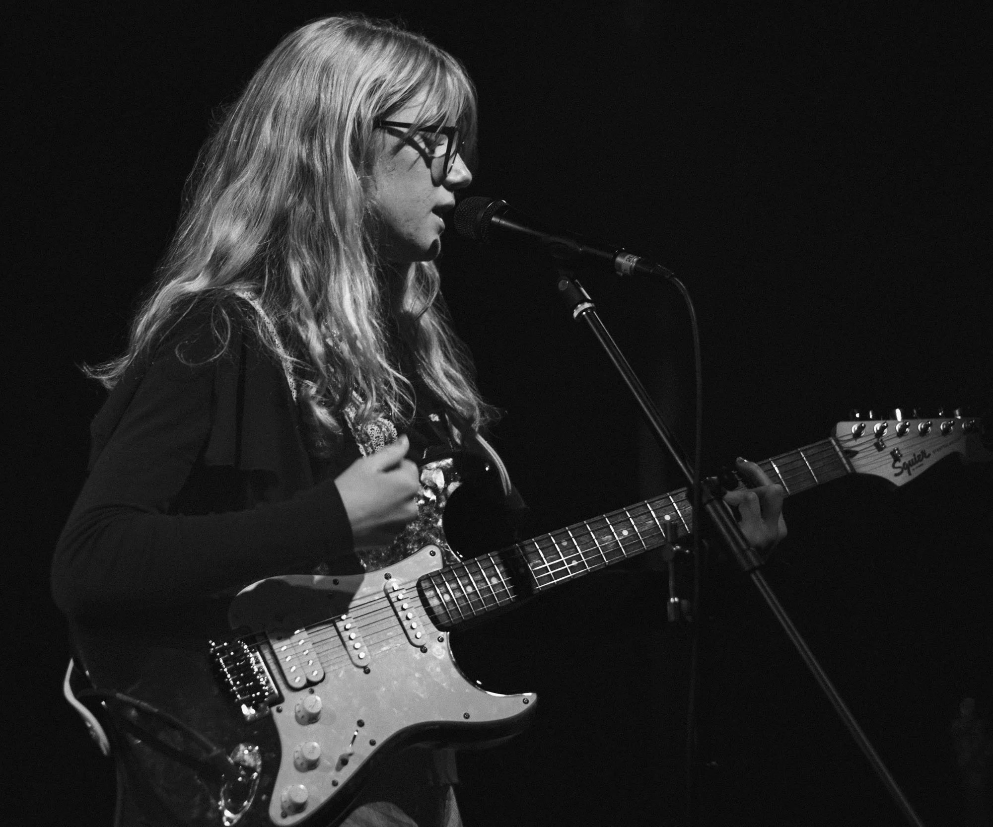 A woman with long wavy hair and glasses playing an electric guitar on stage, singing into a microphone against a dark background.
