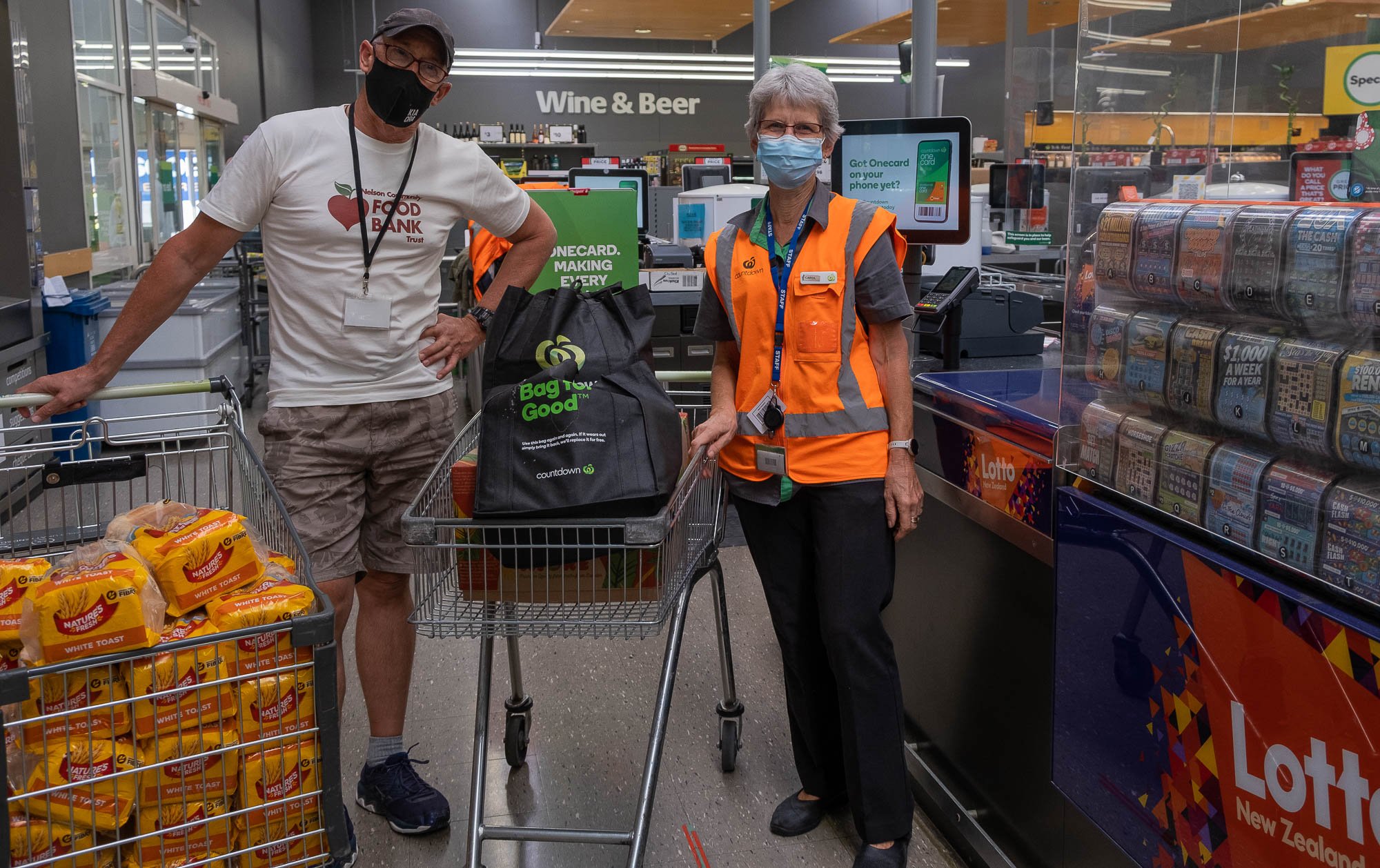 A man and a woman wearing face masks inside a grocery store checkout line. The man, dressed casually, is pushing a shopping cart filled with bread. The woman, wearing an orange safety vest and a store name badge, is standing next to a lotto ticket di