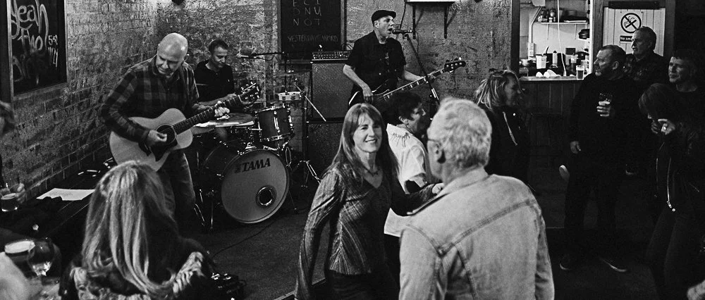A black and white photo of a lively bar with a live band performing. The band consists of a drummer, two guitarists, and a singer. Several patrons are dancing and socializing around the small stage, enjoying drinks and music.