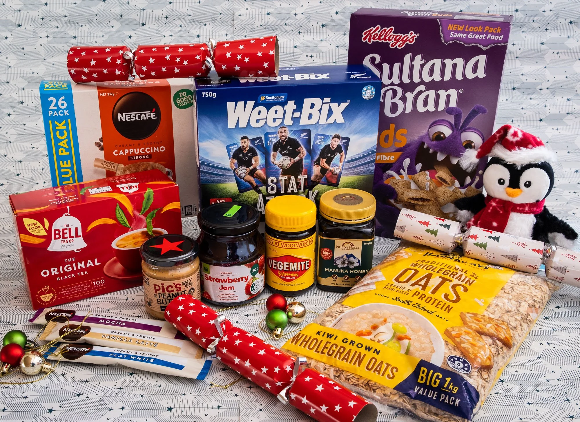 A collection of food and gift items, including boxes of Weet-Bix cereal, Kellogg's Sultanabran cereal, Nescafe cappuccino mix, tea, jam, peanut butter, honey, oats, and decorated festive holiday crackers, with plush penguin and purple monster toys, a