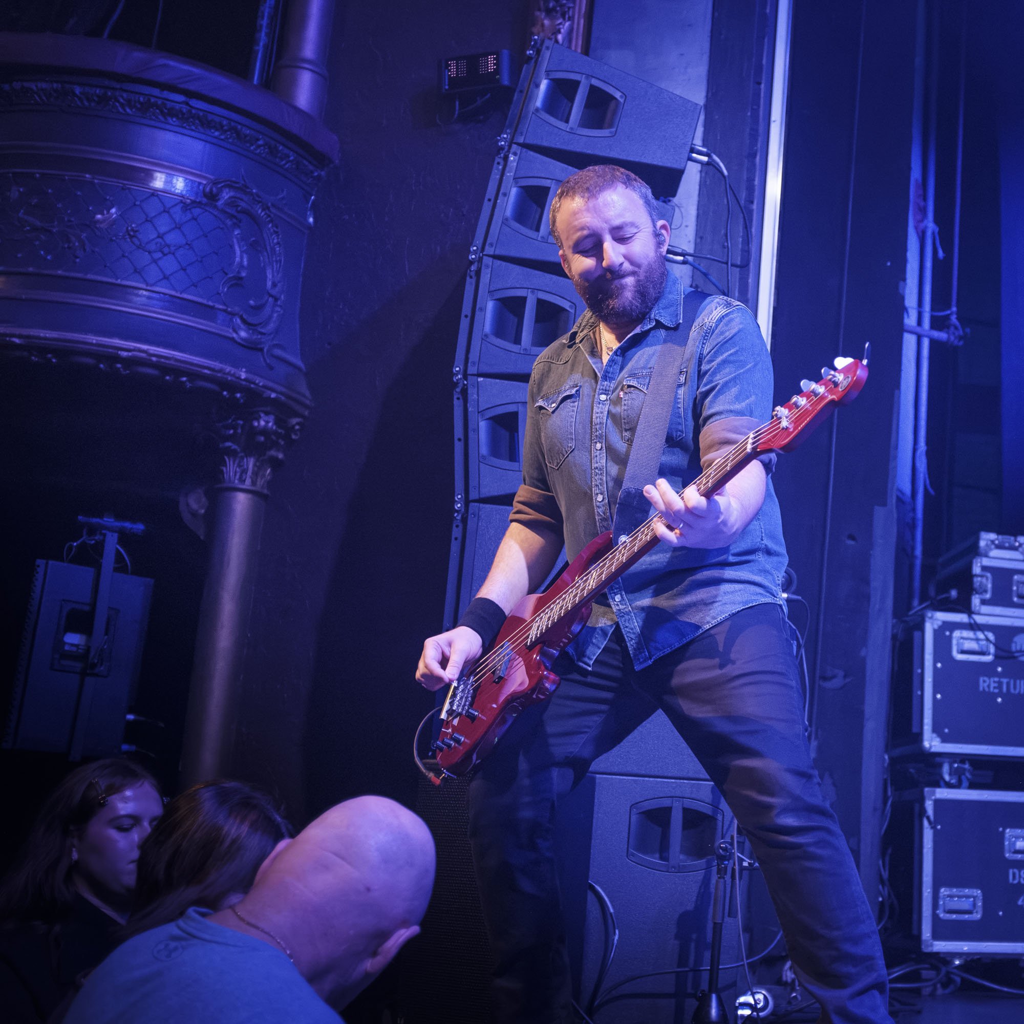 A musician with short hair and a beard playing an electric guitar on stage, with stage equipment and blue lighting in the background, and several audience members watching.