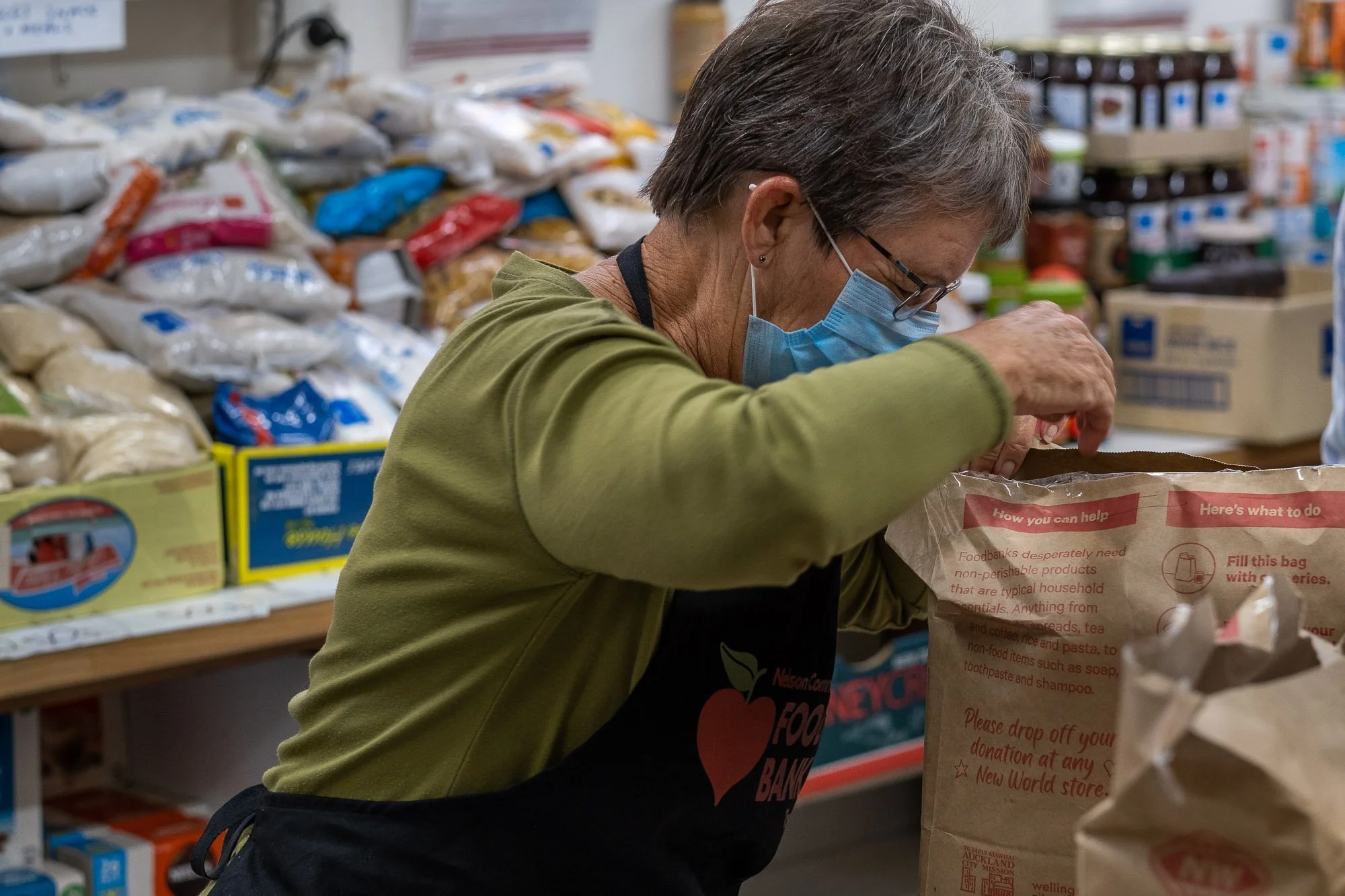 An elderly woman wearing a face mask and glasses is packing food into a paper bag in a food bank. She is wearing a green long-sleeve shirt and an apron that says 'Food Bank'. In the background, there are shelves of packaged food items.