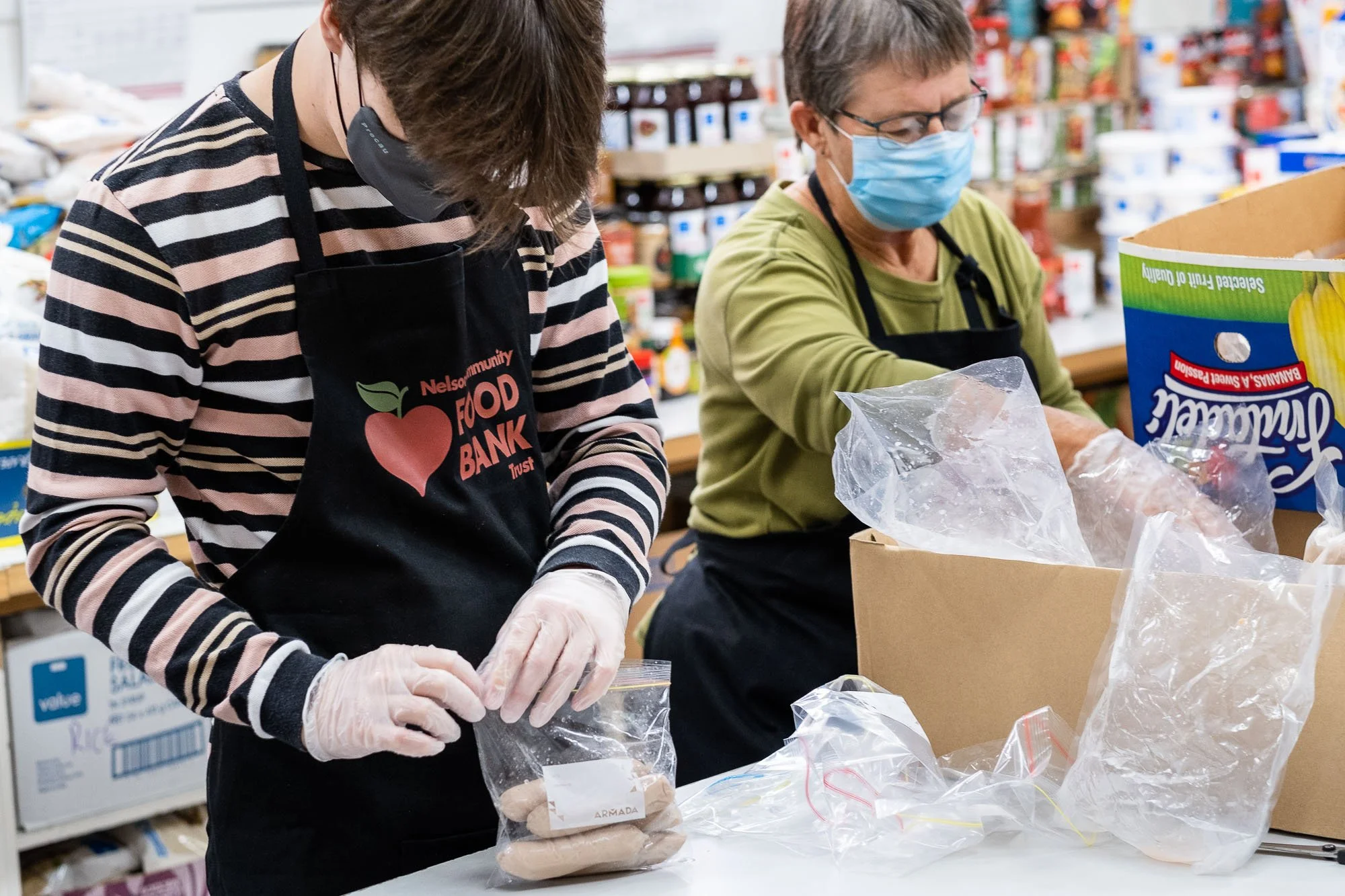Two volunteers wearing masks and gloves packaging food items at a food bank.