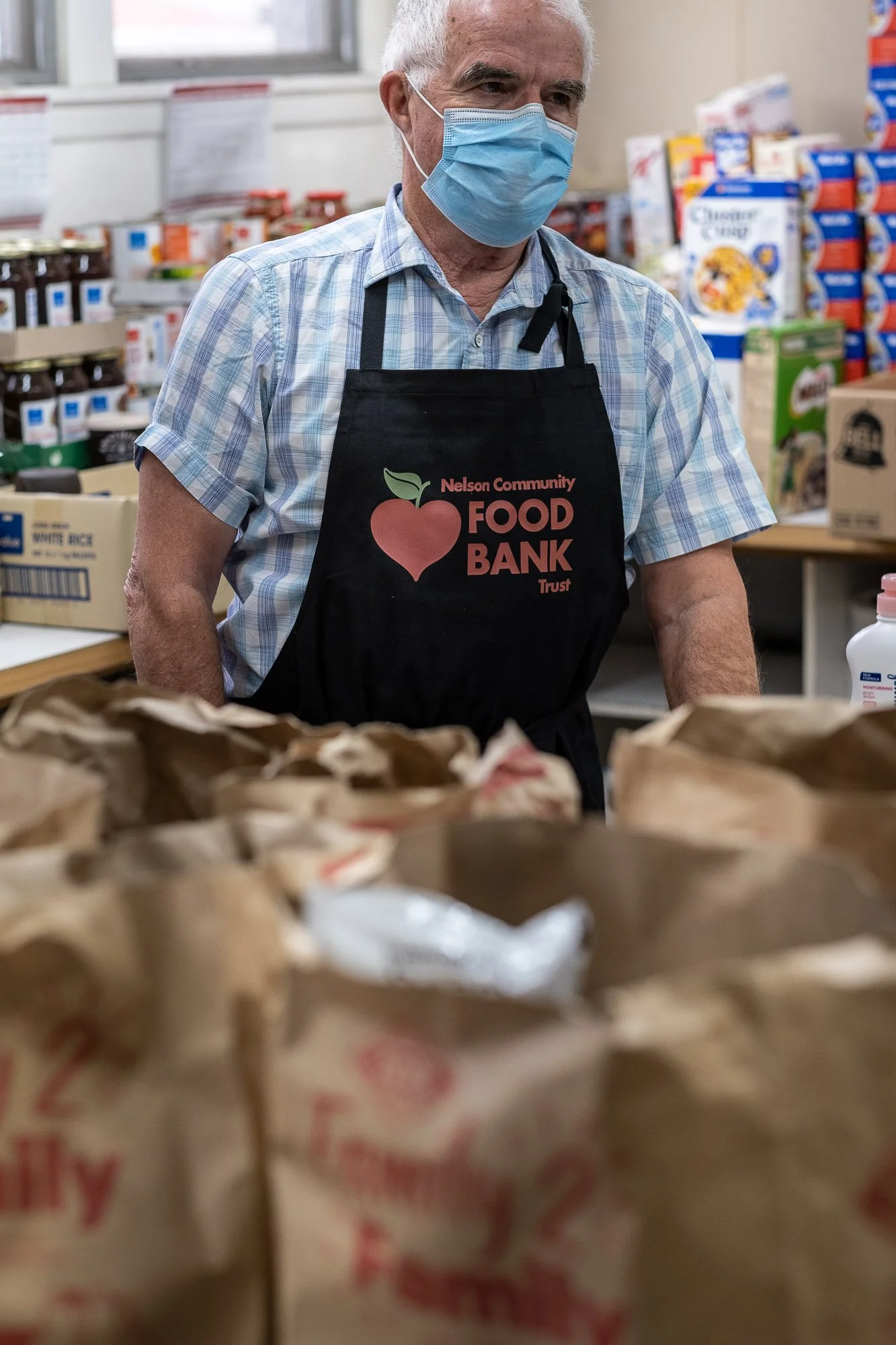 An elderly man wearing a blue face mask and a black apron with an apple logo and the words 'Nelson Community Food Bank Trust' is working in a food bank. The background shows shelves stocked with various food items and boxes.