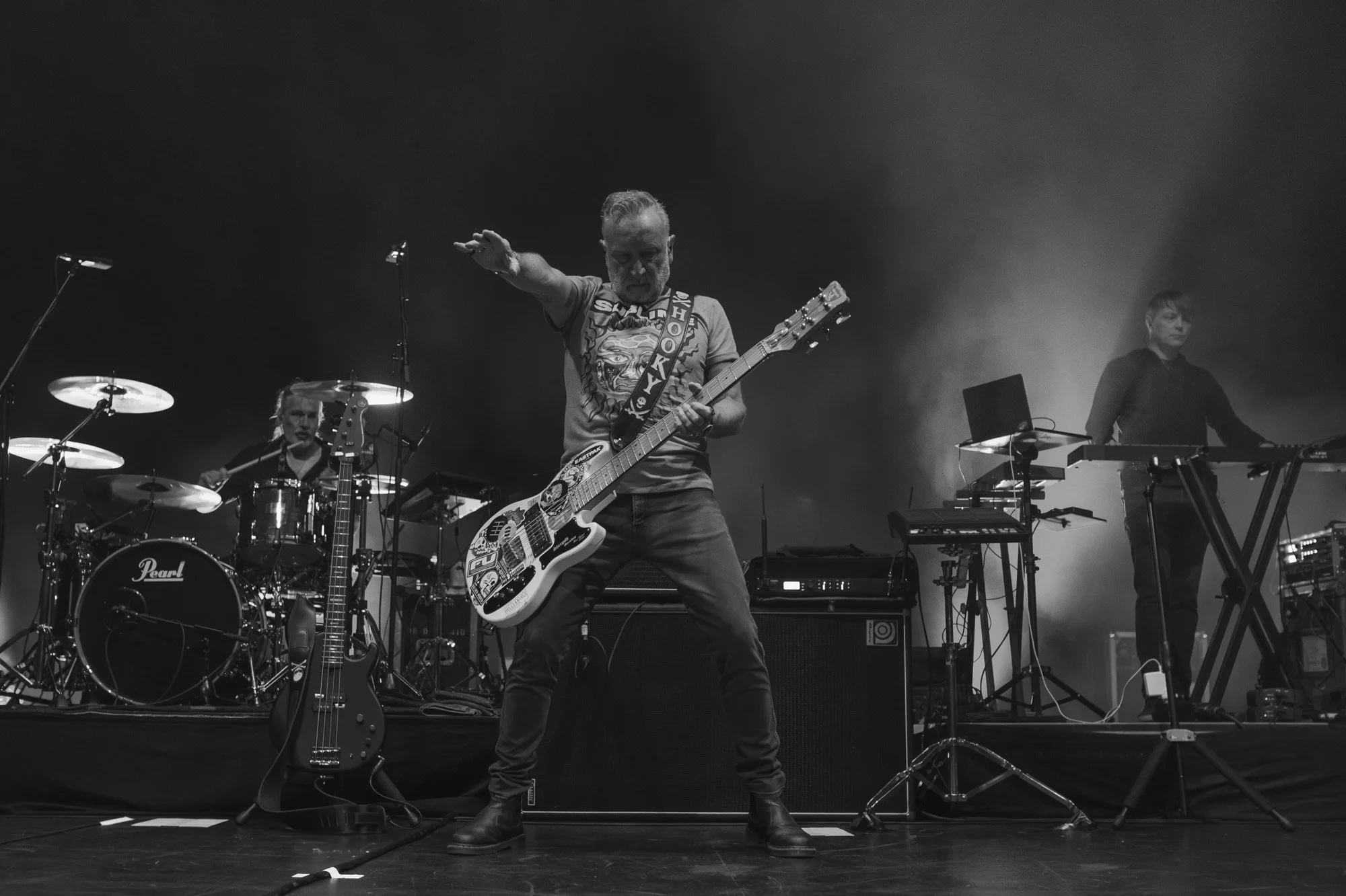 Black and white photo of a band performing on stage. The lead guitarist in the foreground is playing an electric guitar, wearing a graphic T-shirt and jeans, with one arm raised. Behind him, a drummer is behind a drum set, and to the right, another m