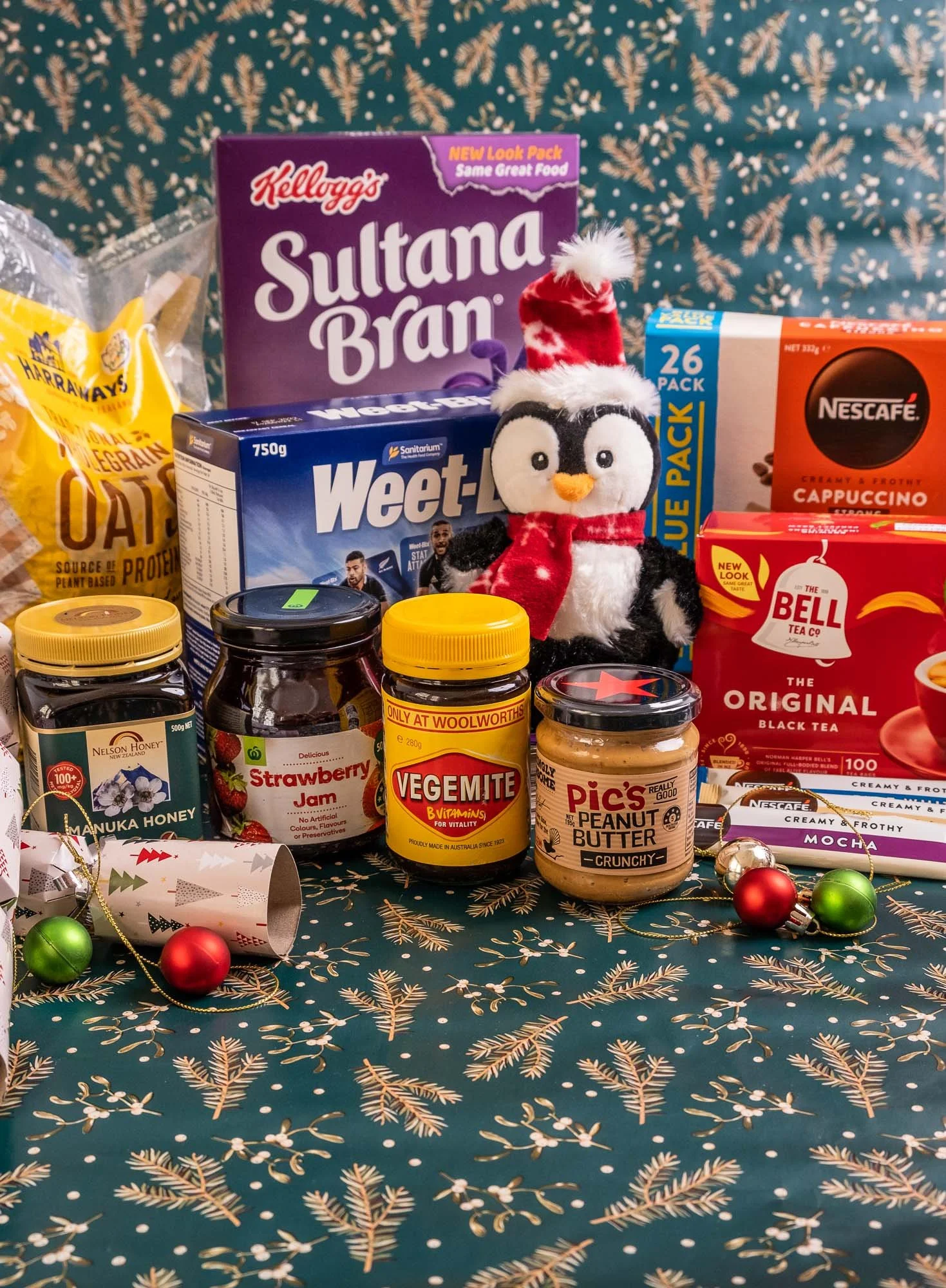 A festive holiday table display with various food products, a plush penguin wearing a Santa hat, and Christmas decorations including small ornaments and a paper party horn, set on a dark green tablecloth with gold pine branch patterns.