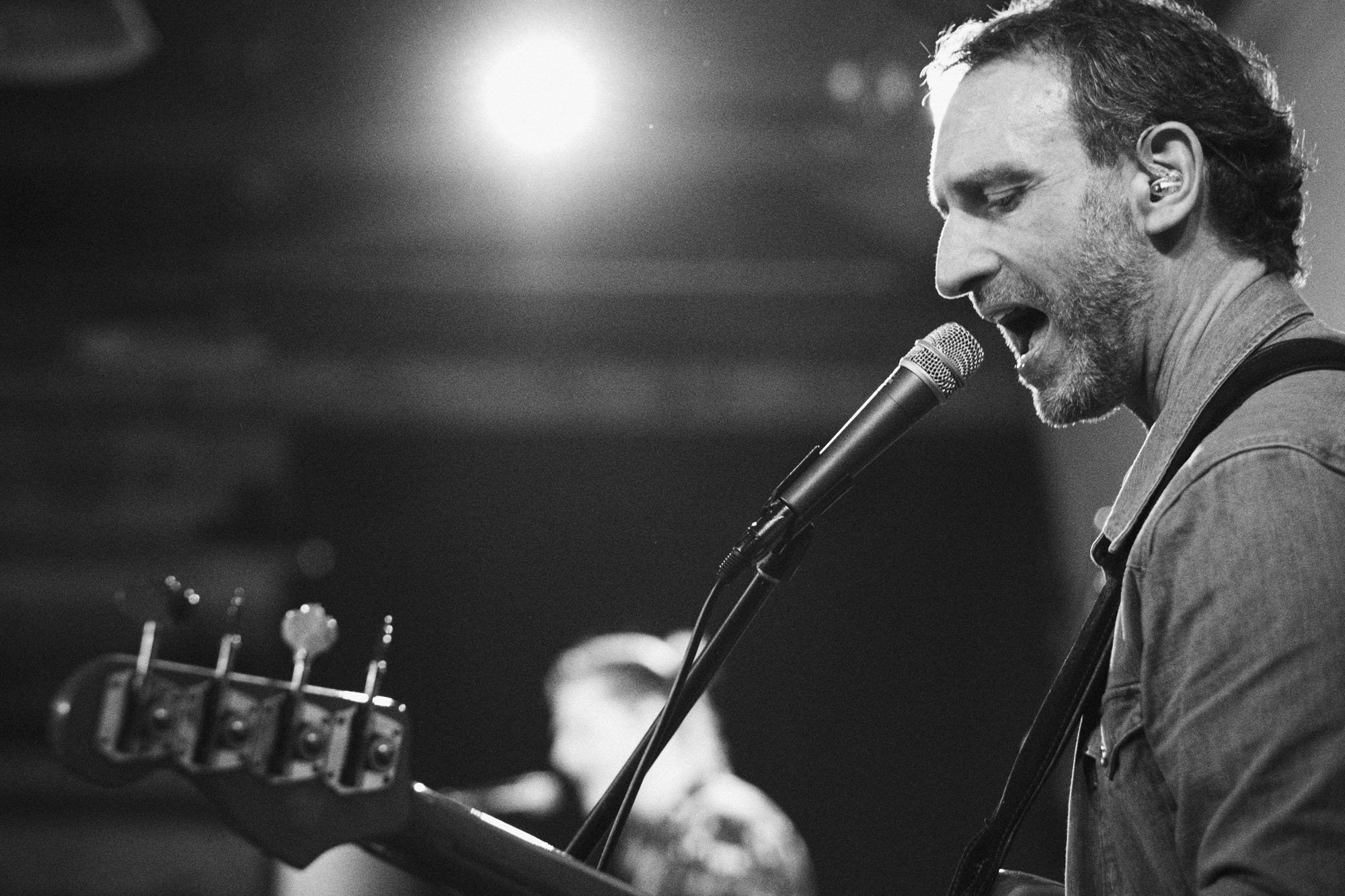 A man with a beard and curly hair singing into a microphone while playing an acoustic guitar on stage in black and white.