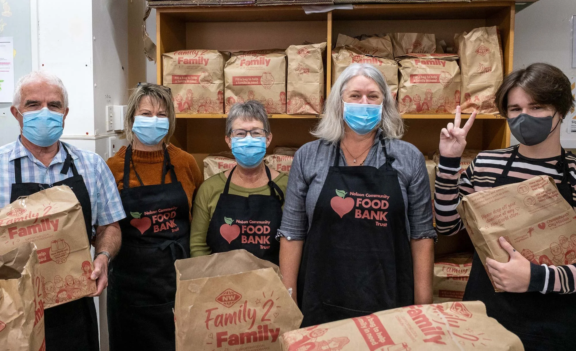 Five people wearing masks and aprons with 'Nelson Community Food Bank' logo standing behind tables filled with paper bags of food, in front of shelves stocked with bags from 'Family' brand.