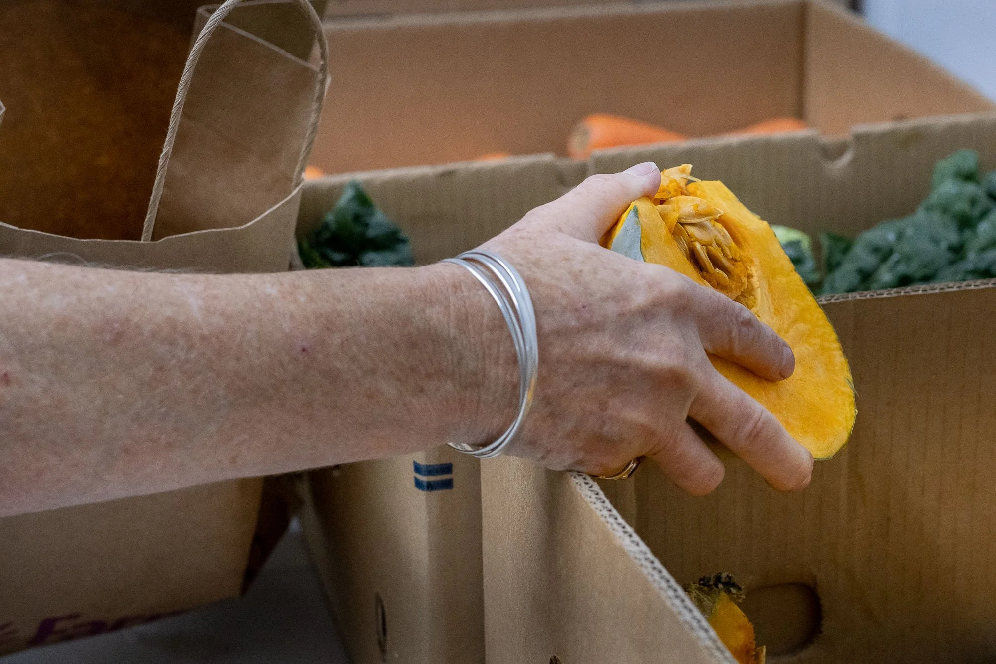A person with a silver bracelet on their wrist is holding a slice of orange pumpkin over a cardboard box filled with green produce.
