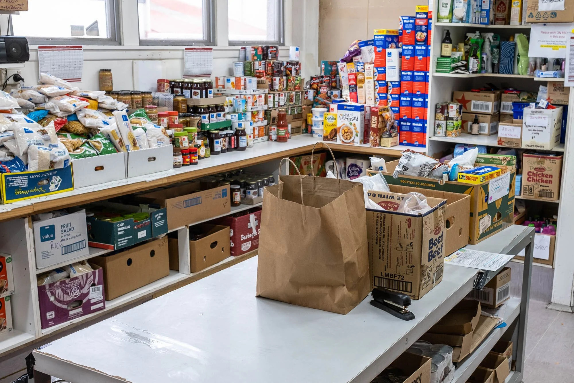Inside a grocery or convenience store with shelves stocked with packaged food, canned goods, and boxes. A table has brown paper bags, a cardboard box, and a stapler on it, with more boxes underneath.