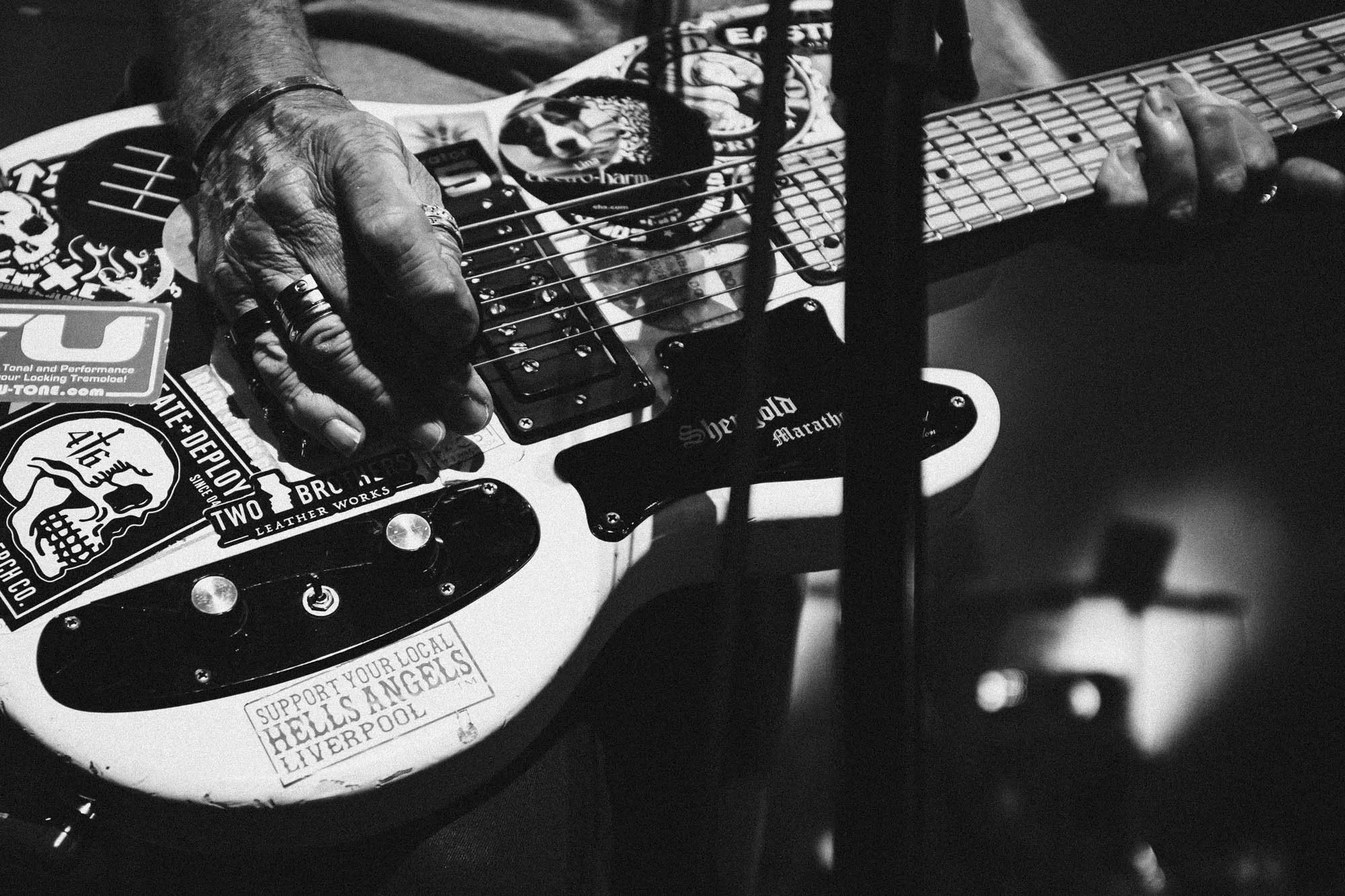 Close-up black and white photograph of an older person's hands playing an electric guitar, decorated with various stickers and patches, on a dark background.