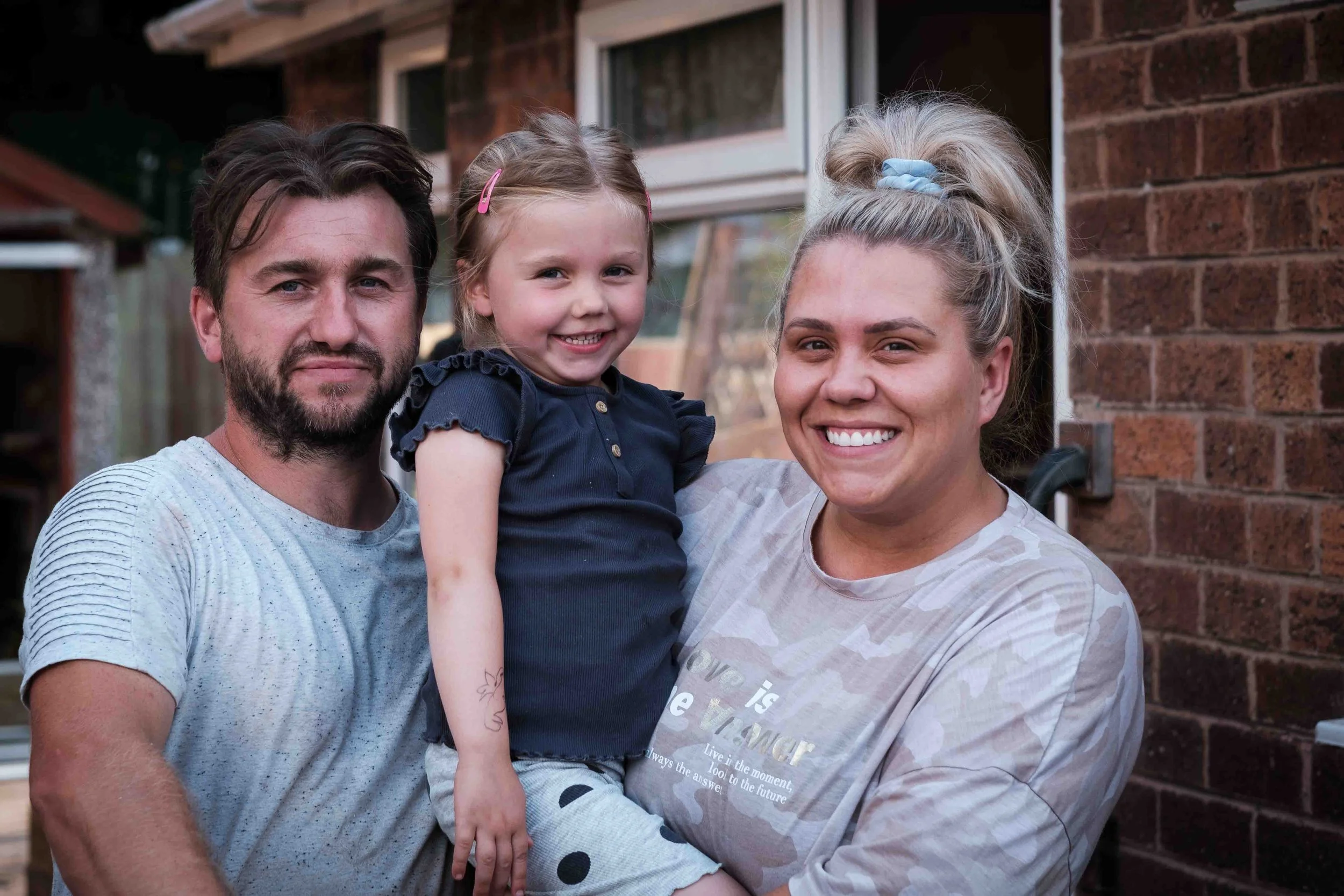 Family of three, father, mother, and daughter, standing outside near a brick house, smiling and looking at the camera.