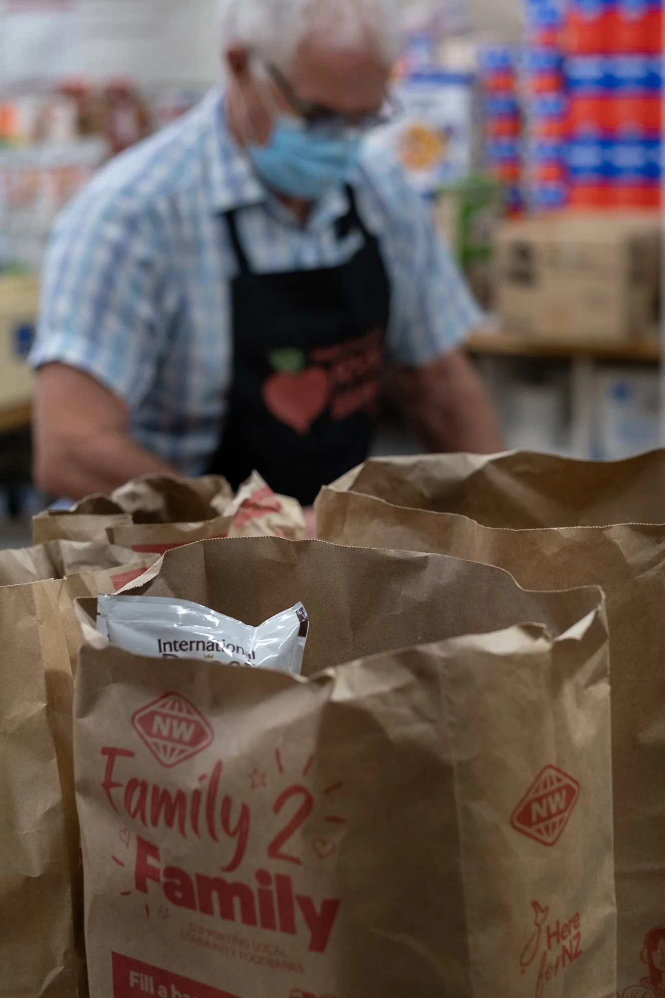 A person wearing a face mask and apron working at a food bank or pantry, with brown paper bags in the foreground labeled "Family 2."
