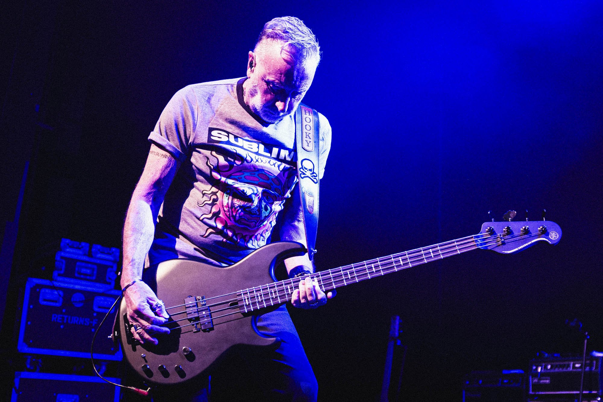A man with gray hair playing a bass guitar on stage under purple and blue lighting, wearing a graphic T-shirt with the words 'SUBLIM' and a skull design.