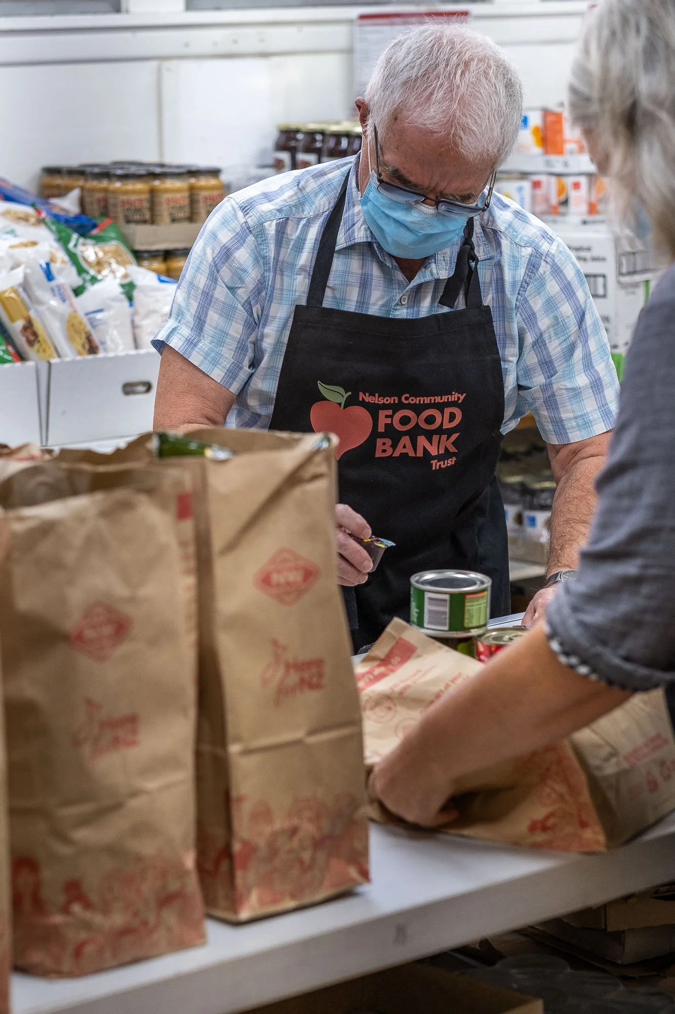 Volunteer wearing a face mask and apron working at a food bank sorting canned and packaged food items on a table.