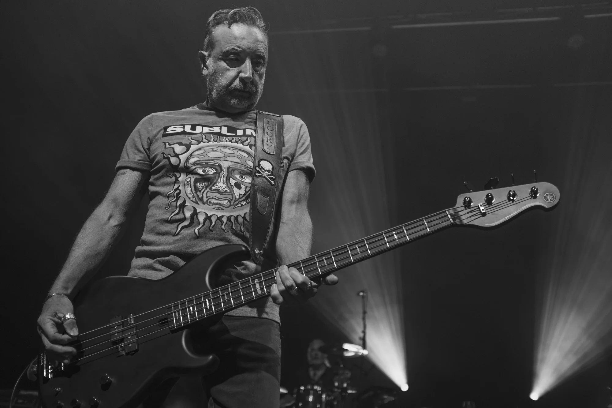 A black and white photograph of a middle-aged man playing an electric bass guitar on stage. He has short, wavy hair and a beard. He is wearing a t-shirt with a graphic print and a guitar strap with a skull and crossbones label. The background is dark