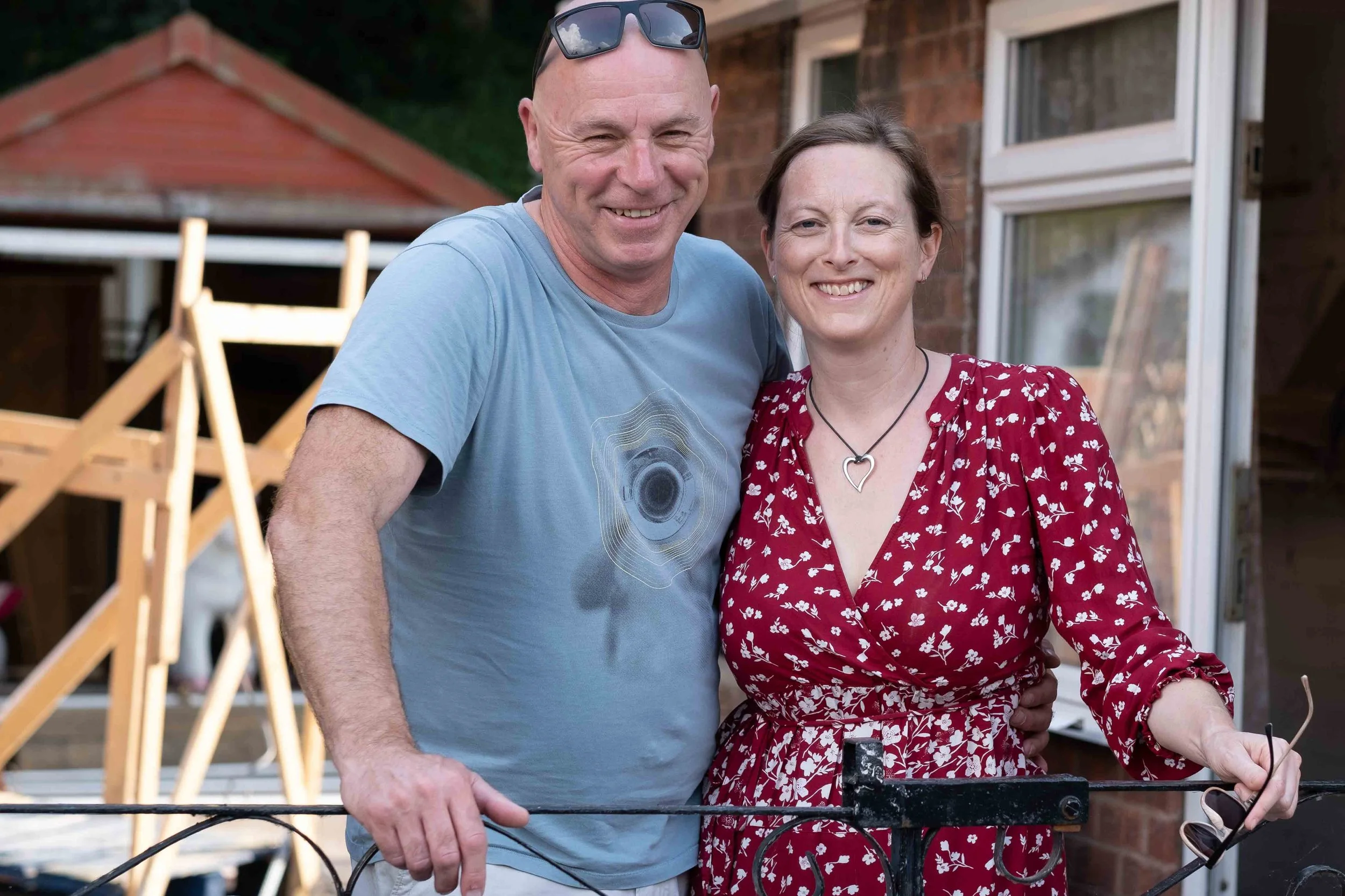 A smiling man and woman standing outdoors near a brick house, embracing each other, with sunglasses on the man's head and her holding a pair of glasses.