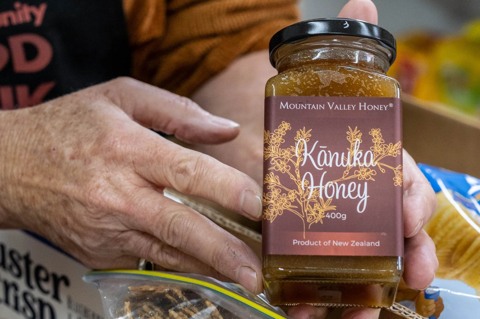 A person's hand holding a jar of Mountain Valley Honey, labeled as Kanuka Honey from New Zealand, with the hand resting near some packaged snacks.