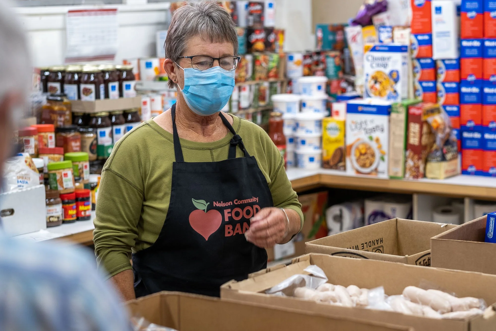 A woman wearing glasses, a face mask, and an apron with 'Nelson Community Food Bank' logo stands behind shopping carts filled with food, in a grocery store or food bank setting with shelves stocked with various food products.
