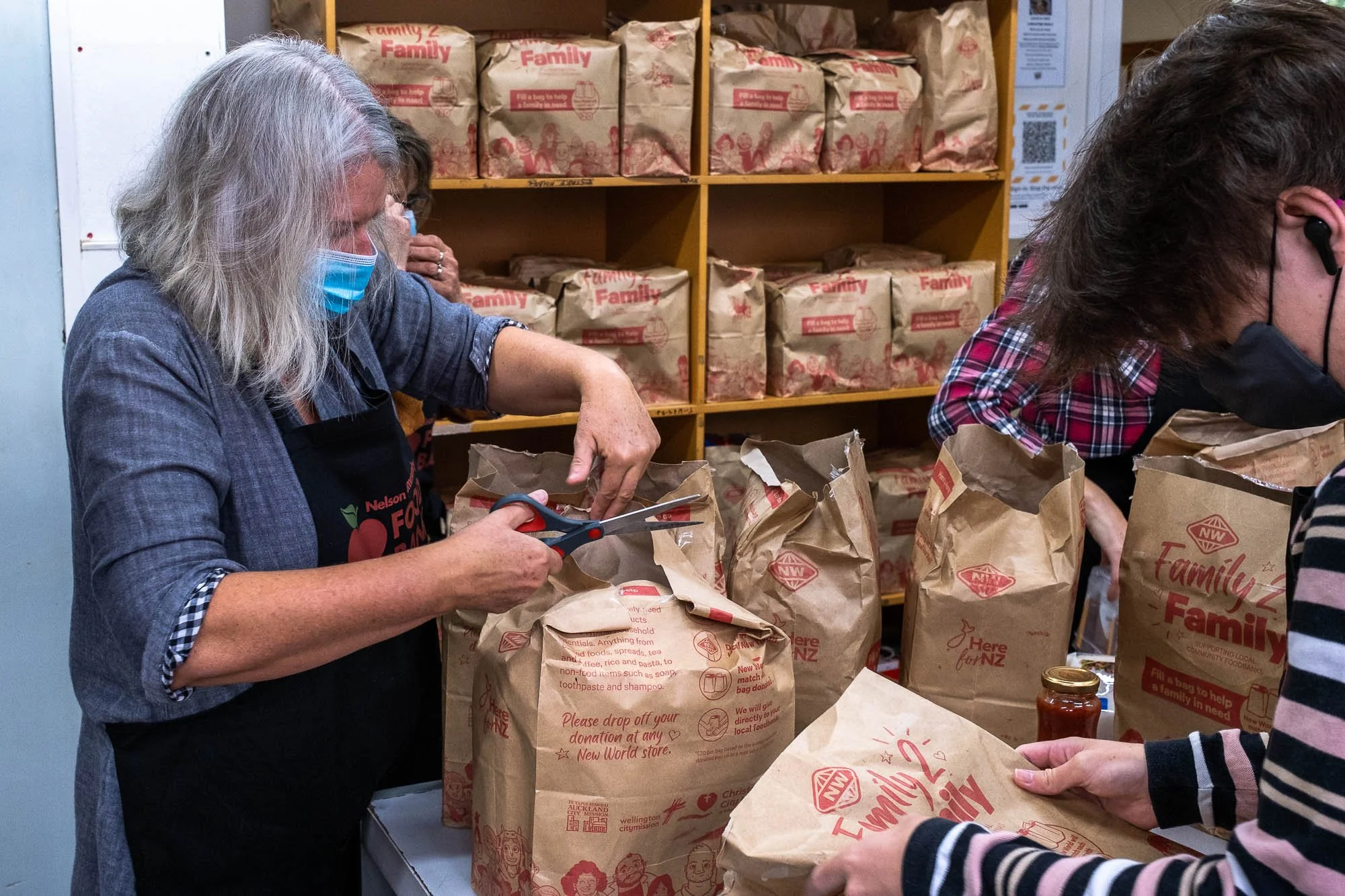 People packing food bags in a community service setting with shelves of supplies in the background.