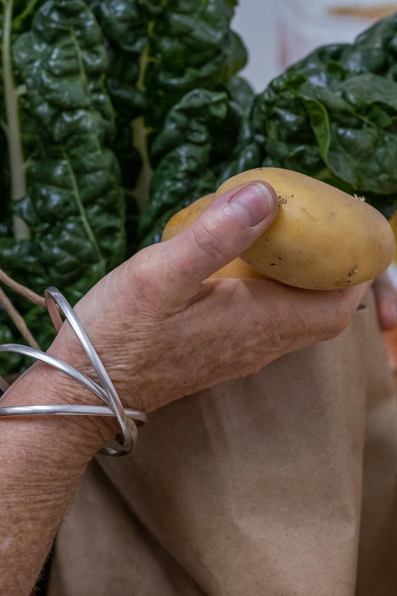 A person's hand holding a yellow potato with lush green leafy vegetables in the background.