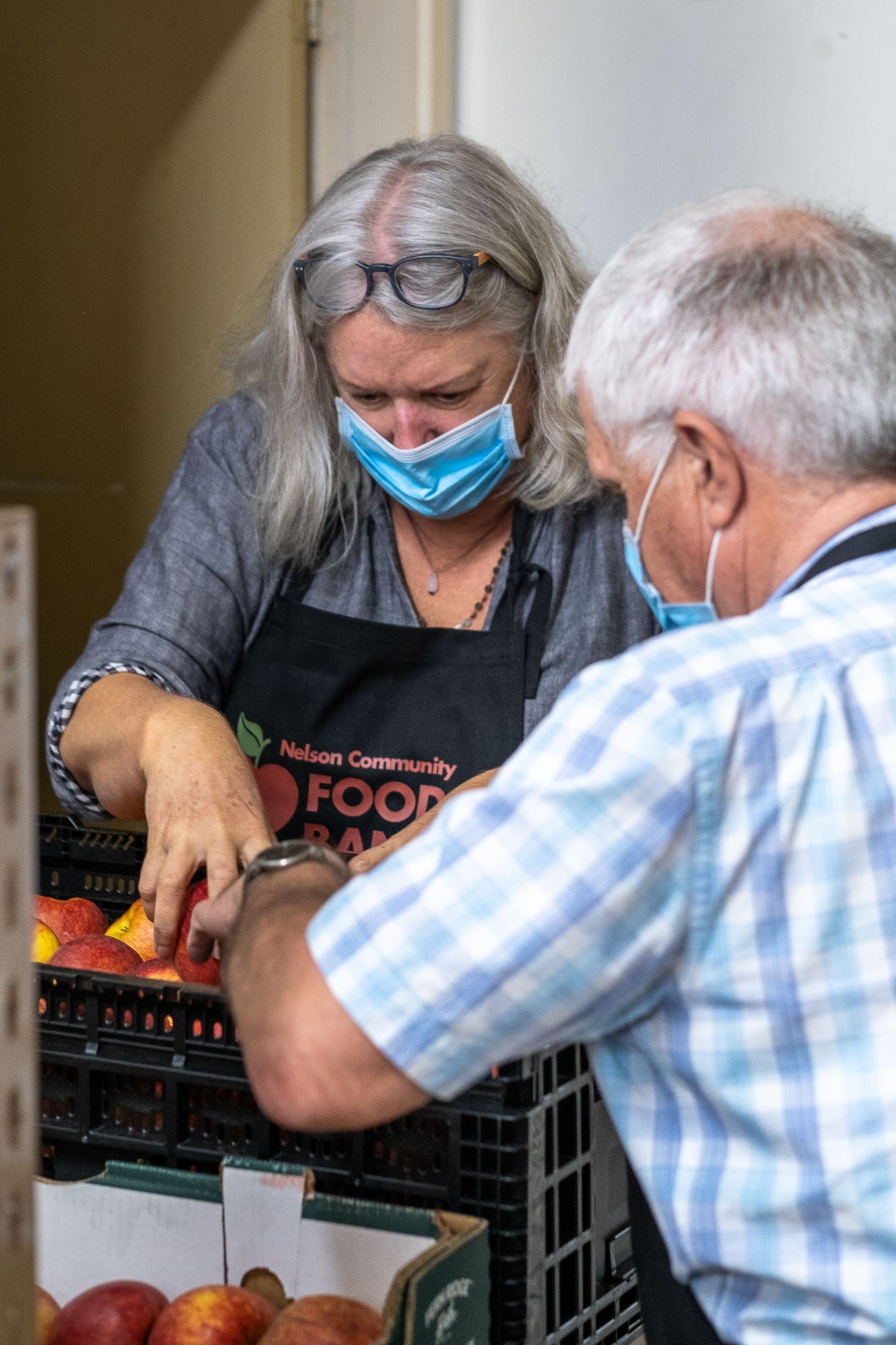 Two people wearing masks and aprons are working together to pack apples at a food bank.