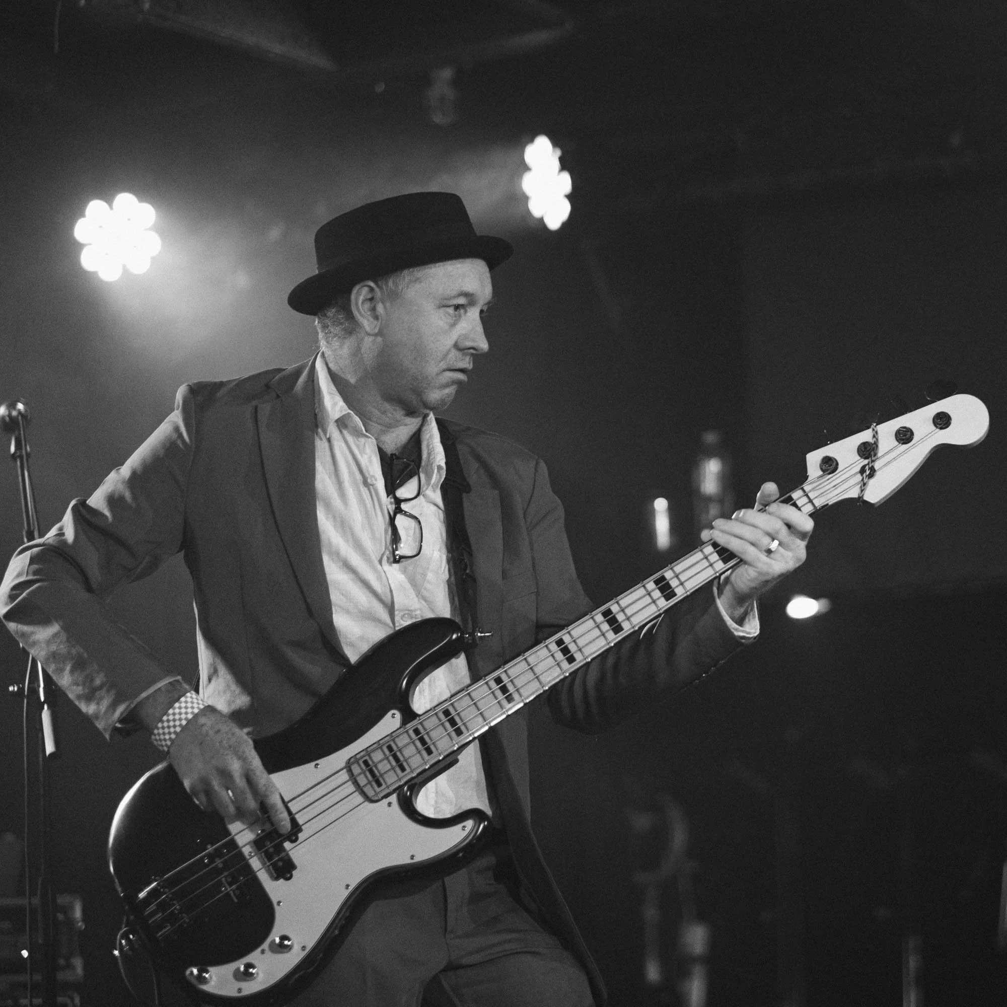 Black and white image of a man wearing a hat and suit, playing an electric bass guitar on stage with blurred bright lights in the background.