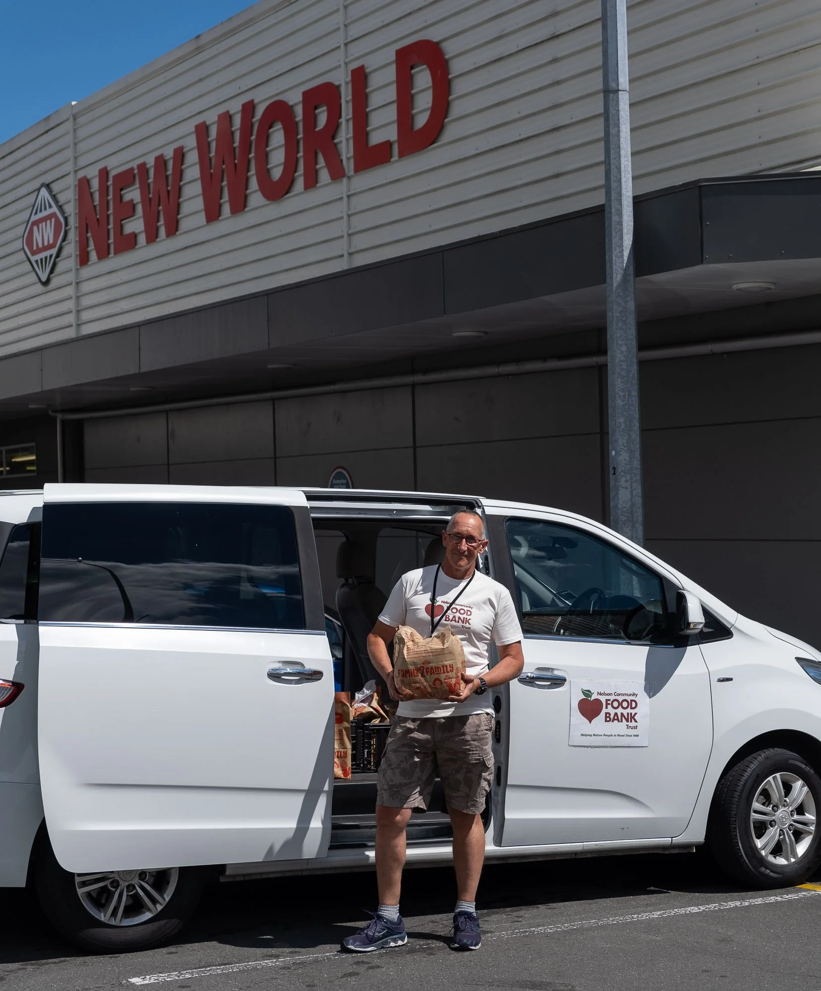 A man standing beside an open car door in a parking lot, holding a paper bag. The car has a sign that reads 'Nelson Community Food Bank Trust.' A large store sign for 'New World' is visible in the background.