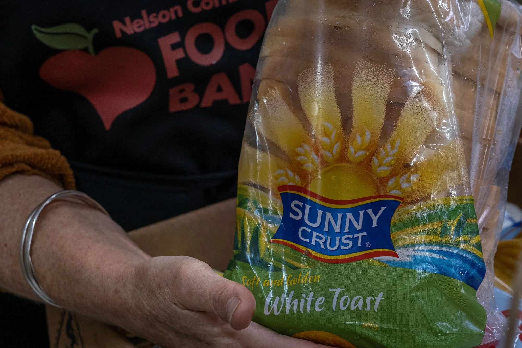 Close-up of a hand holding a bag of Sunny Crust white bread slices with the logo and text visible, and a black shirt with red and green text in the background.