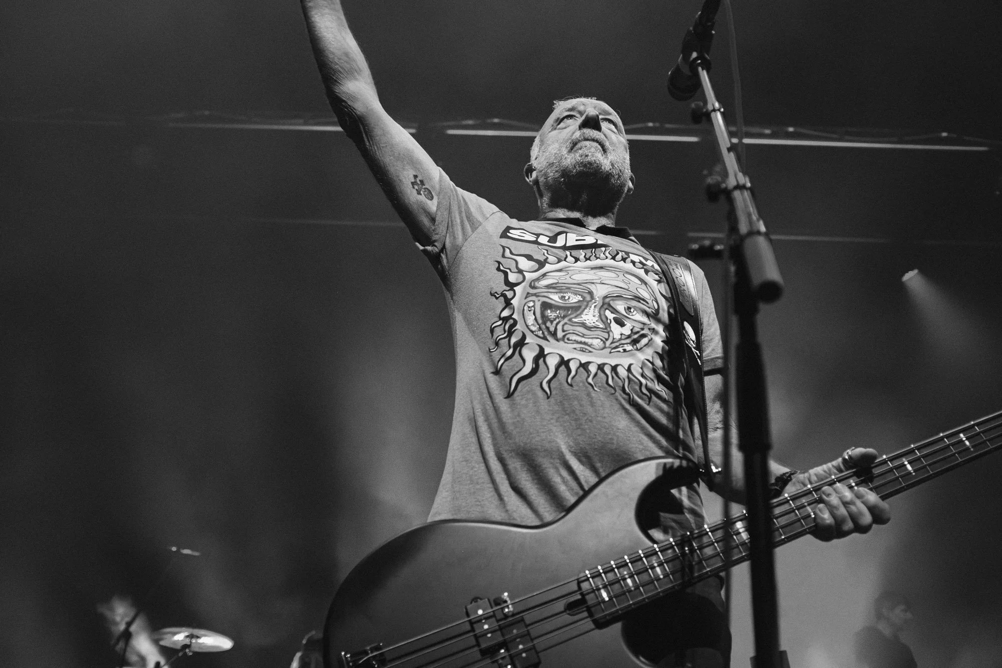 A man with glasses and a beard playing an acoustic guitar on stage, raising one arm, wearing a graphic t-shirt, in black and white.