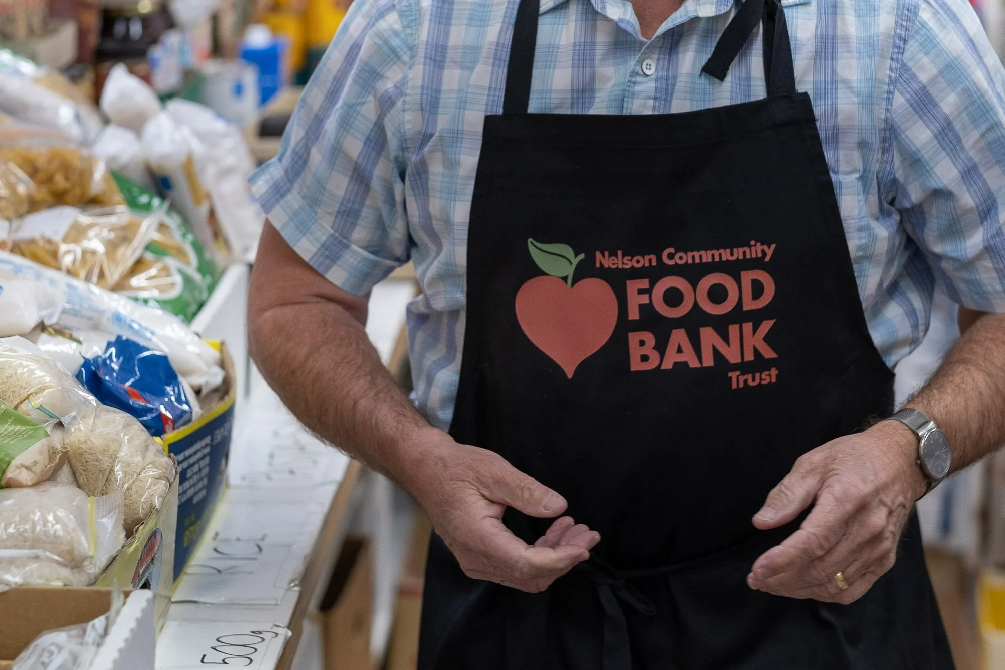 Man wearing a black apron with 'Nelson Community Food Bank Trust' written on it, standing at a food bank with shelves of food packages in the background.
