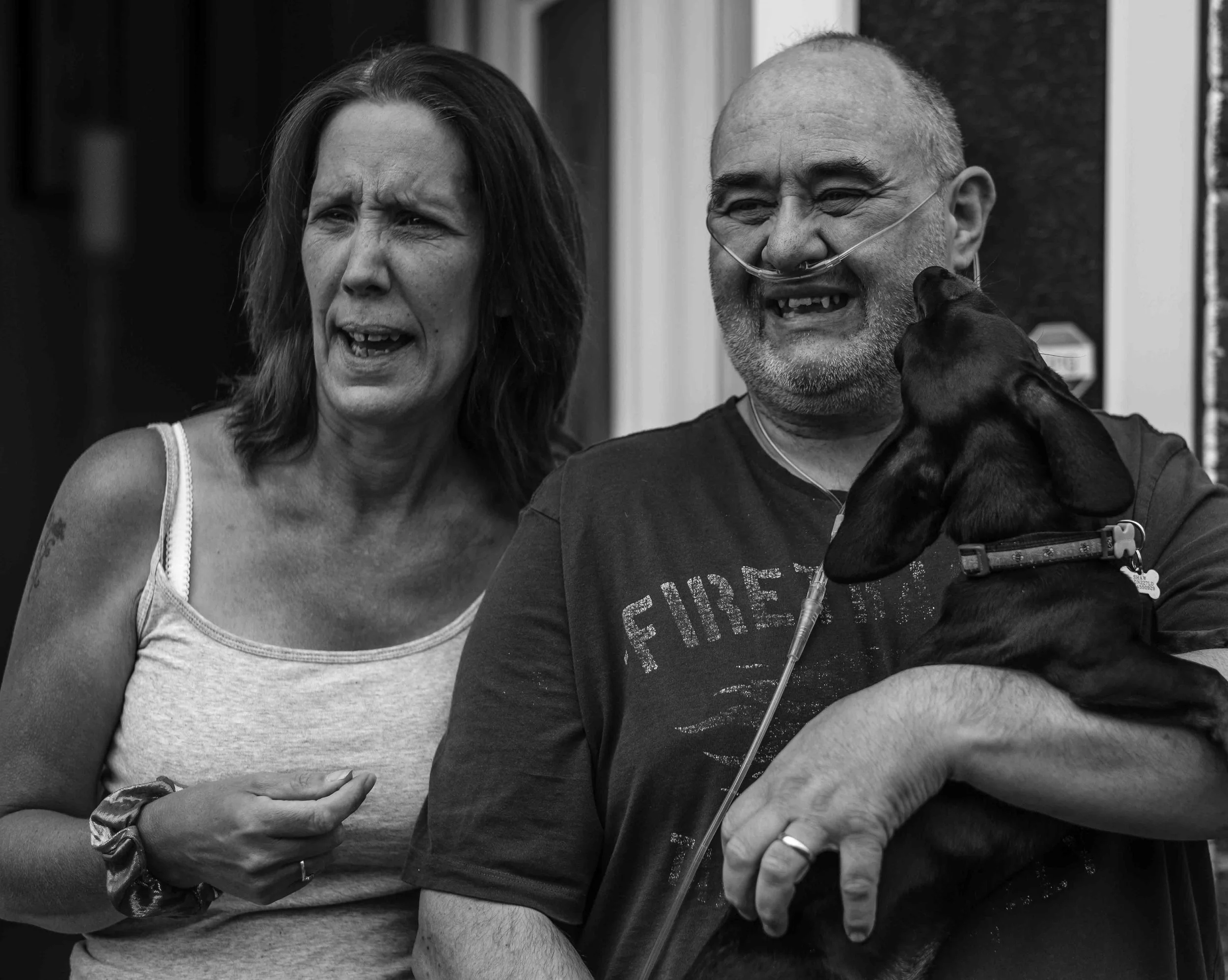A black and white photo of a woman and a man with medical equipment, holding a dog, both appearing emotional.