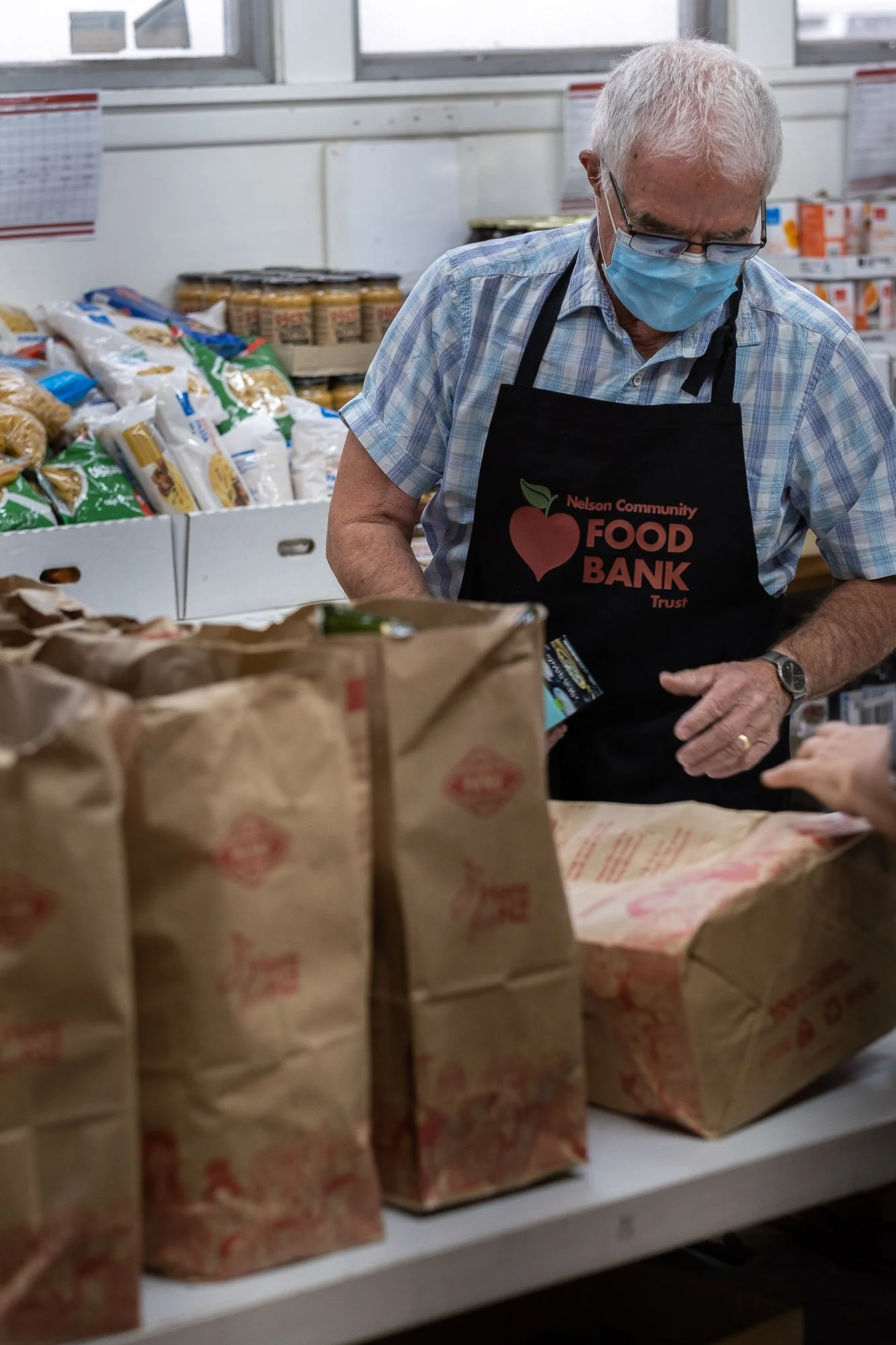 An elderly man wearing glasses, a blue checkered shirt, and a face mask, working at a food bank, sorting food packages. He is wearing a black apron with the text 'Nelson Community Food Bank' and a peach logo. Food supplies are on the table and shelve