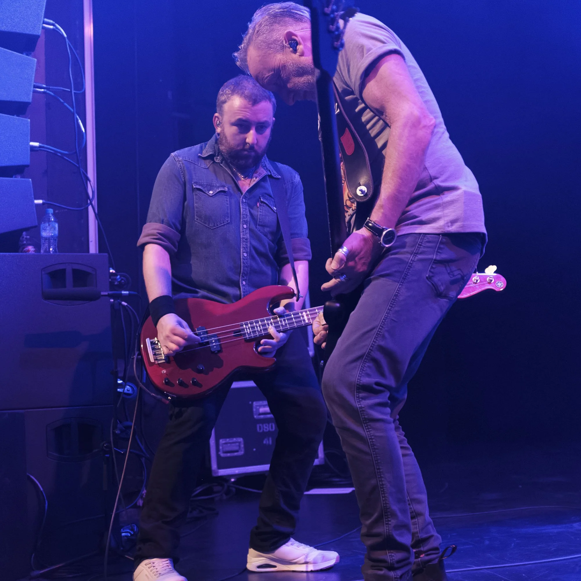 Two musicians on stage, one playing a red electric guitar and the other leaning over him, with stage equipment and purple lighting in the background.