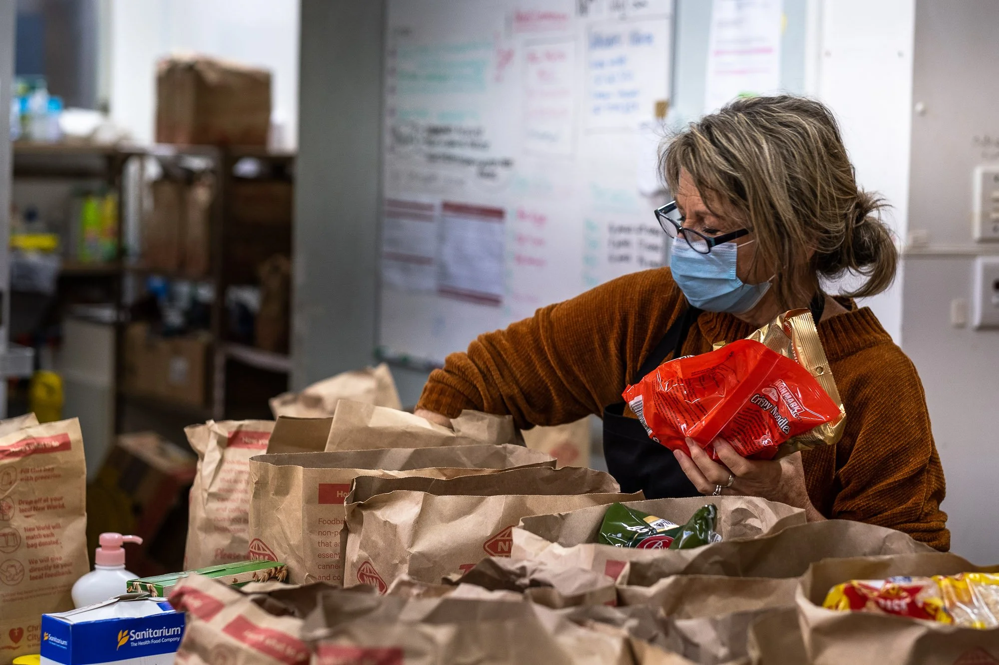 A woman wearing a face mask and glasses sorts groceries at a food bank. She has shoulder-length hair, wears a brown sweater, and holds a red bag of noodles. There are paper bags and various food items on the table in front of her.