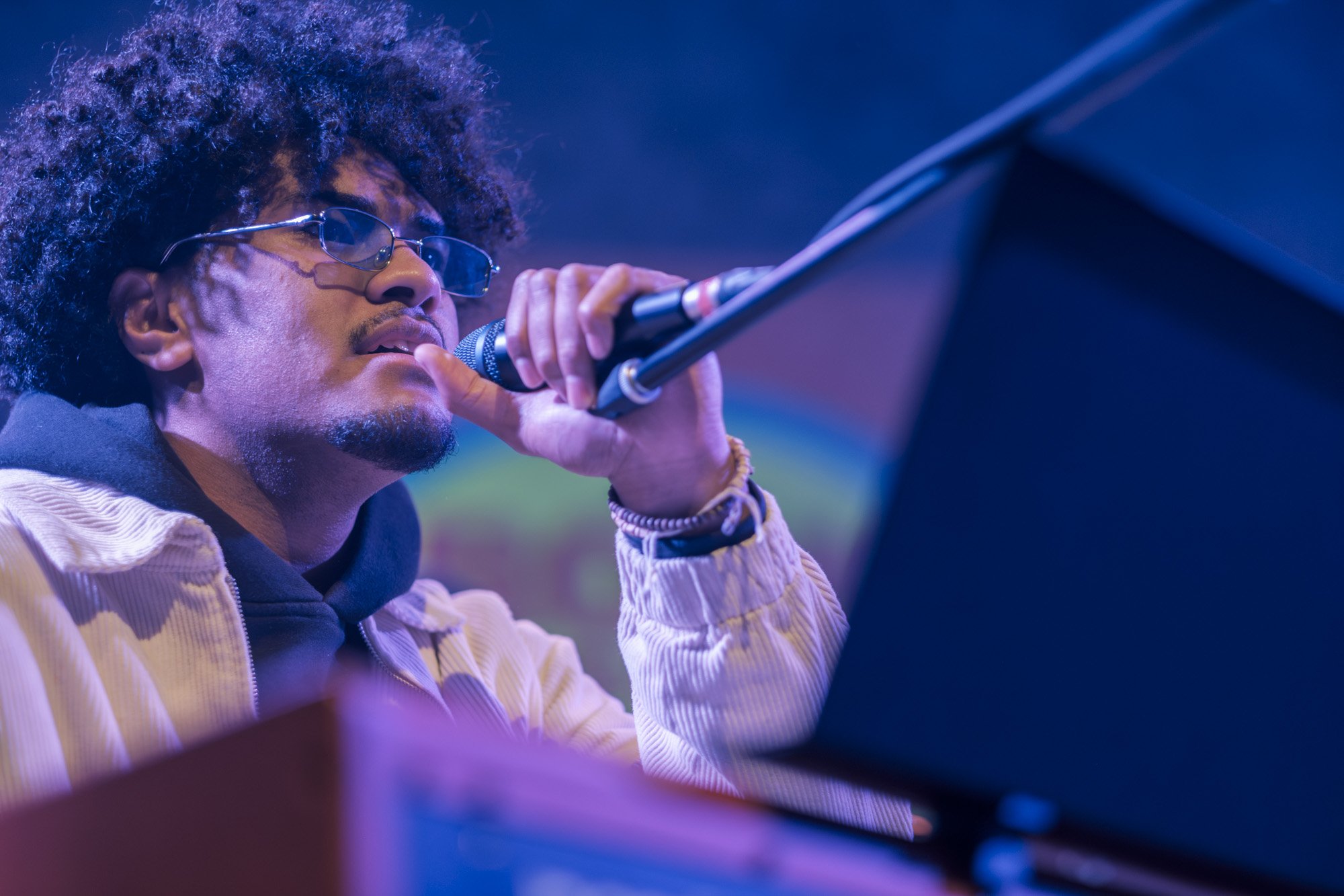 A man singing or speaking into a microphone, wearing sunglasses and a yellow jacket, with a blurred blue and purple background.