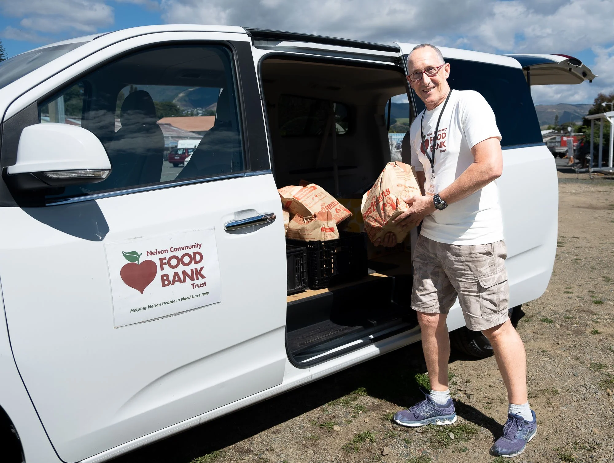 A man wearing a white t-shirt, cargo shorts, and sneakers standing next to a white van with an open side door, unloading paper bags from the Nelson Community Food Bank. The van has a logo with a red apple and green leaf, and the background shows a pa